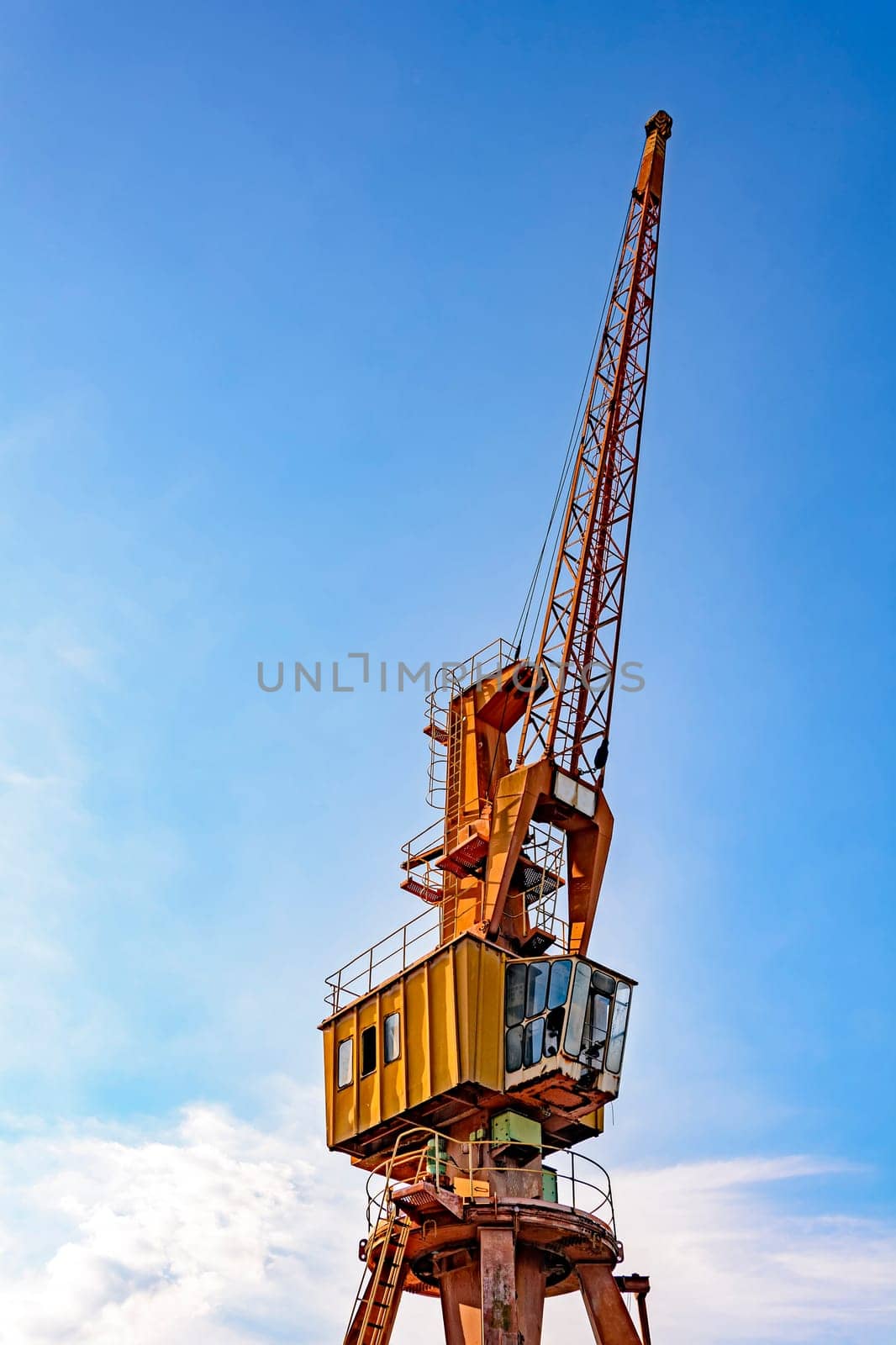 Old and obsolete yellow crane on the harbor pier with blue sky and clouds in the background.