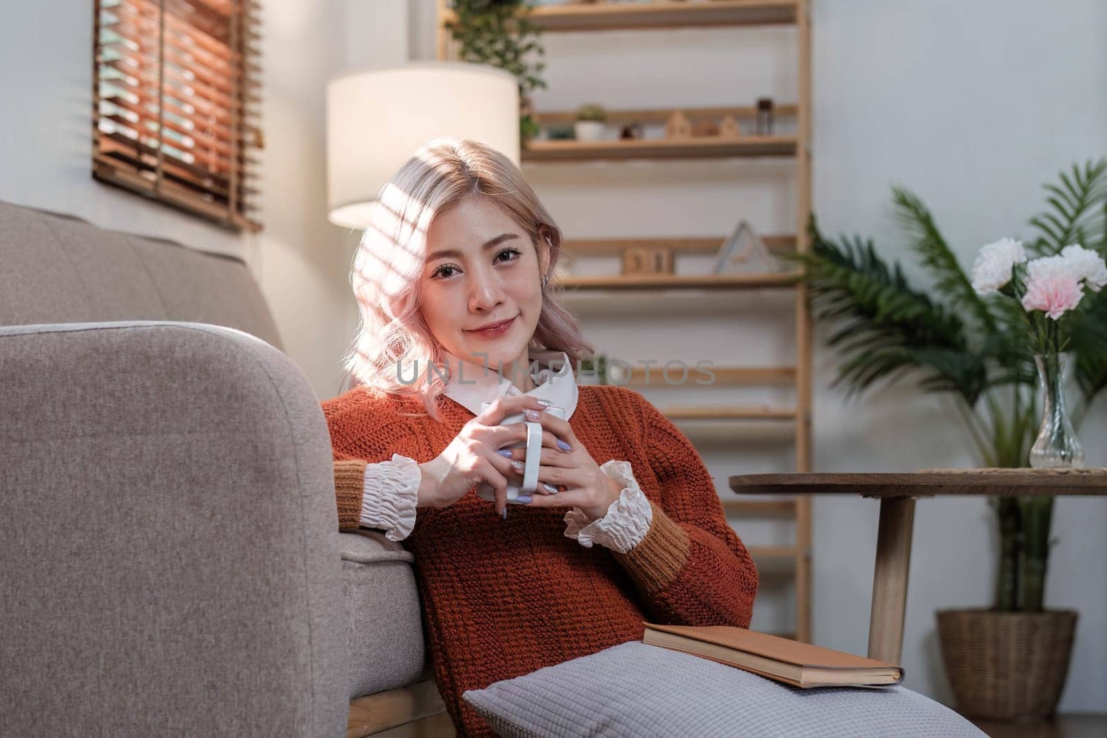 Young beautiful Asian woman relaxing in living room at home, reading books, drinking coffee and using phone in the morning with happy feeling..