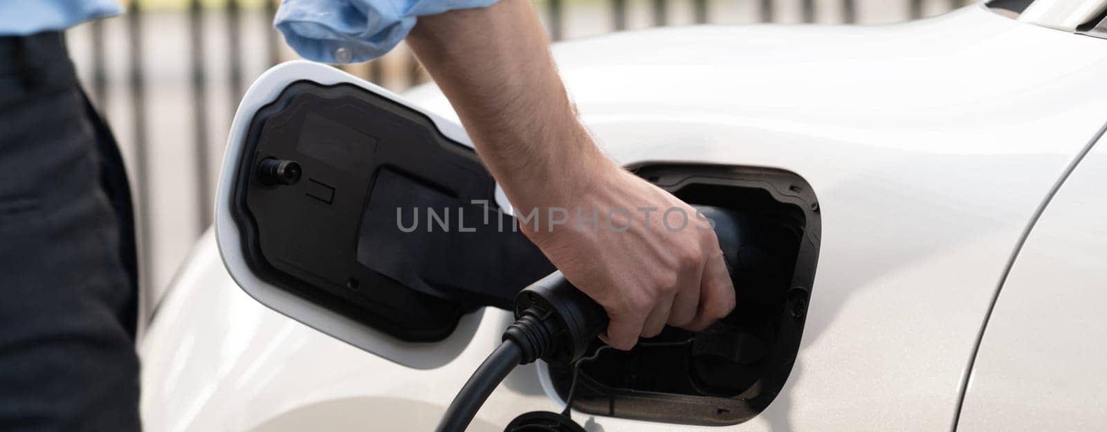 Closeup progressive man holding EV charger plug from public charging station for electric vehicle with background of residential building as concept eco-friendly sustainability energy car concept.