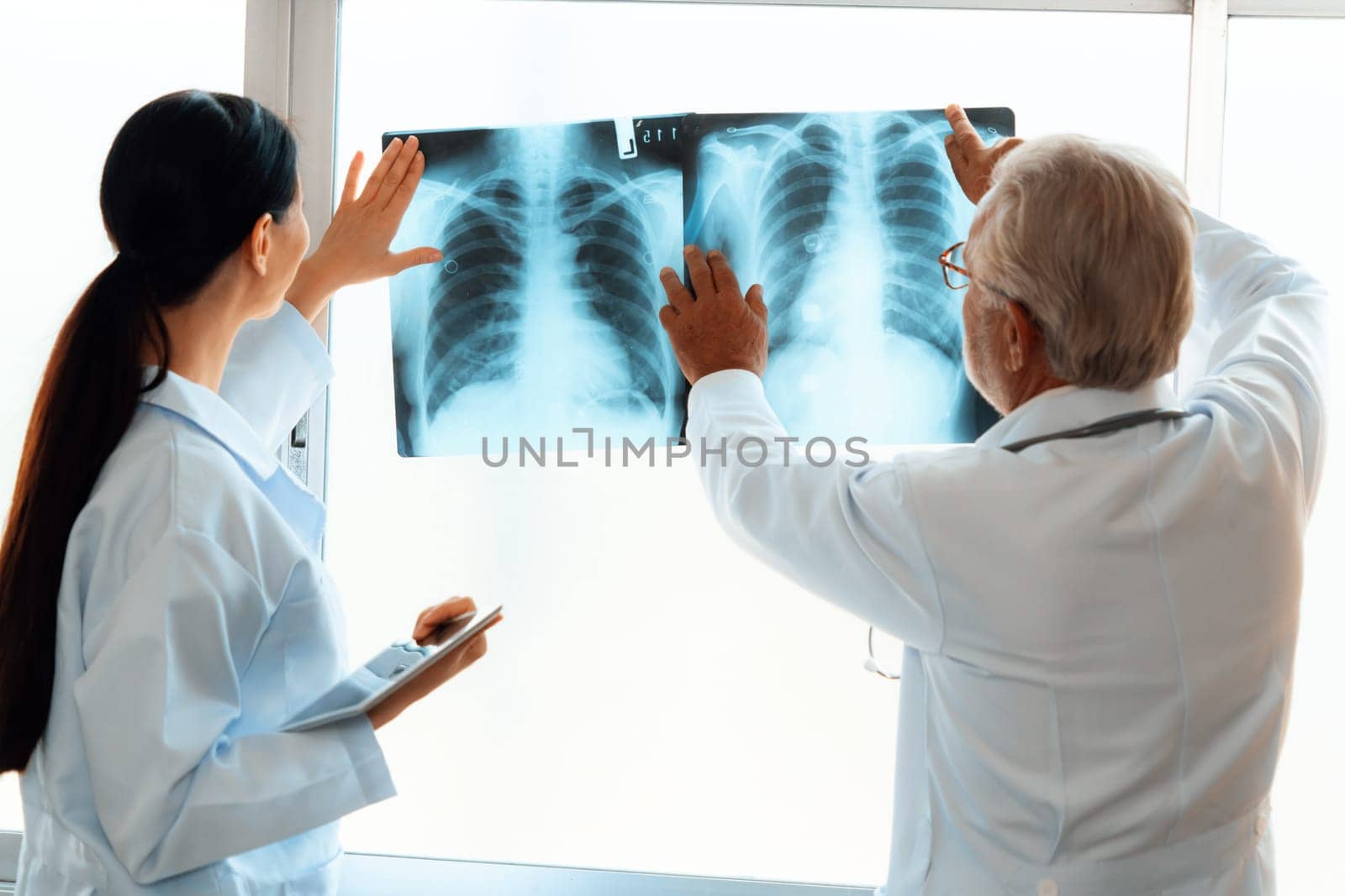 In a hospital sterile room, two professional radiographers hold and examine a radiograph for medical xray diagnosis. Novice doctor seeks advice on a patient's condition from experienced older doctor.