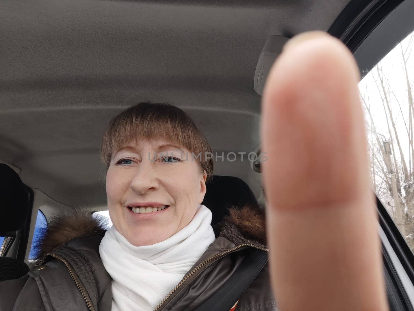 Portrait of adult cute funny blonde woman in warm clothes taking a selfie inside the car and driving it. The concept of pleasant trip in spring, autumn, winter