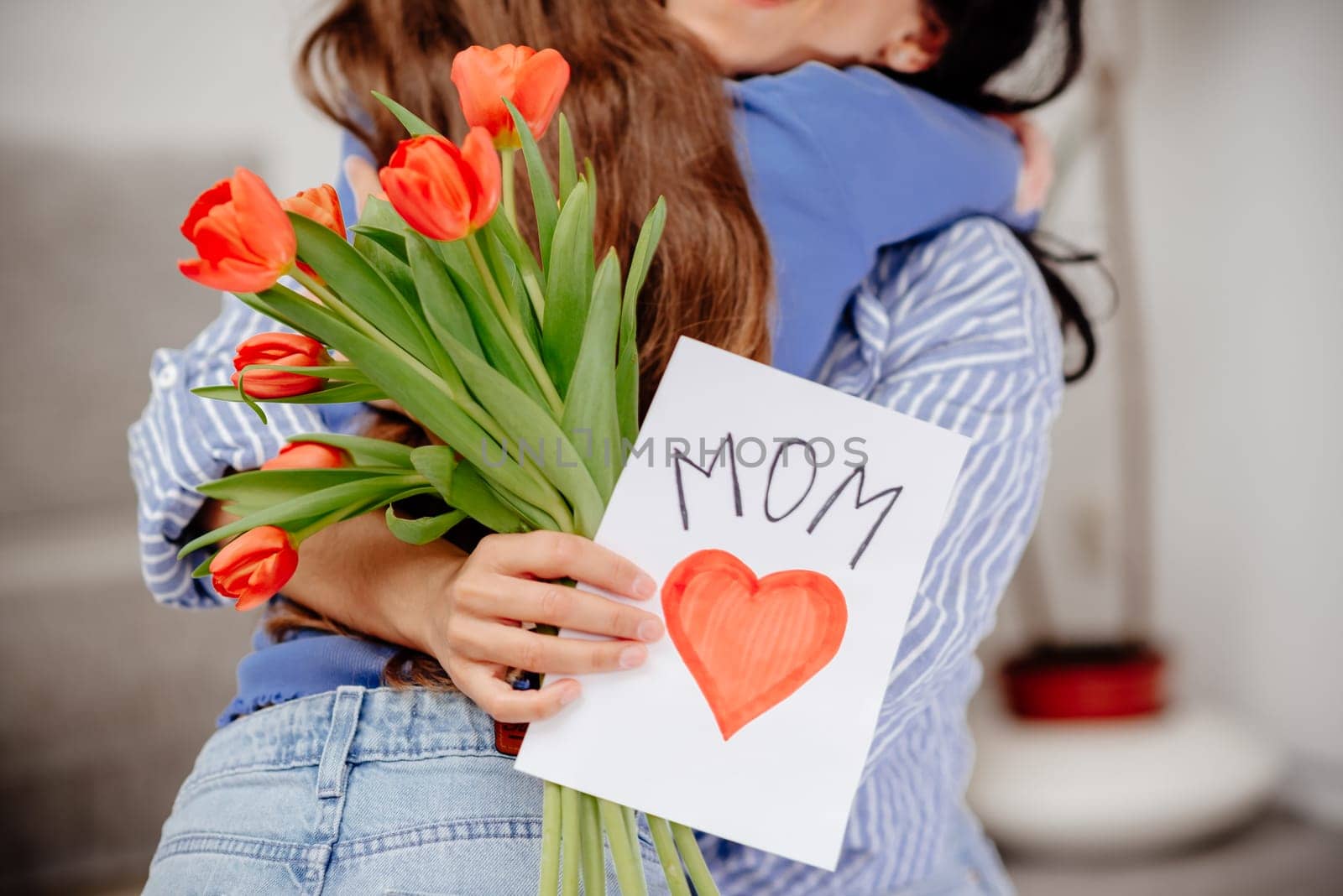 Grateful mom hugging daughter girl, holding flowers bouquet, receiving hand drawn greeting card with loving heart from girl, smiling at camera. Mothers day, 8 march, concept. Head shot portrait. Close-up of hands holding a bouquet of tulips and a handmade card.