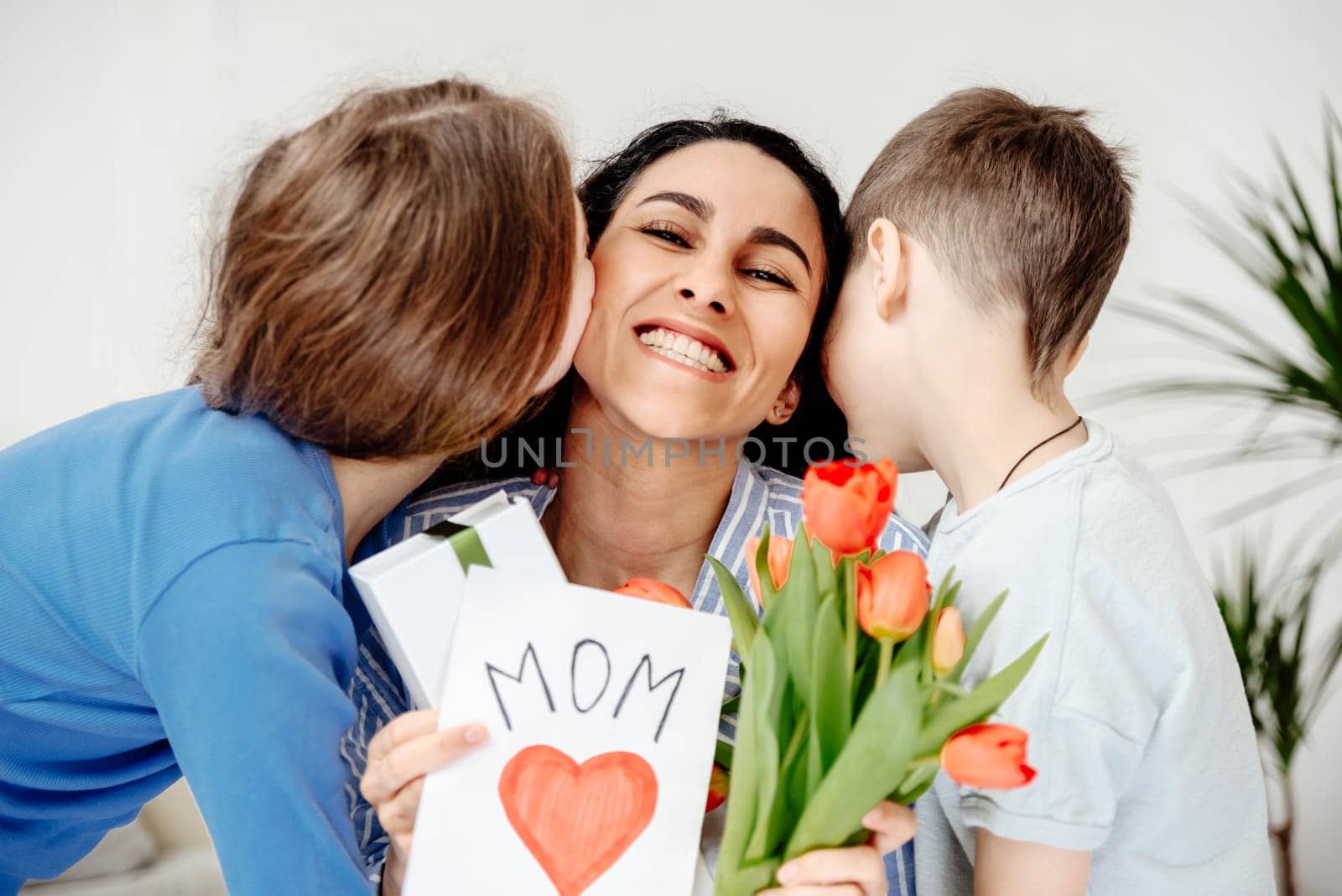 The children congratulated their mother on Mother's Day, gave her a bouquet and a handmade card. A heart is drawn on the card and the word mom is written. Son and daughter pressed cheek to mother's cheek.