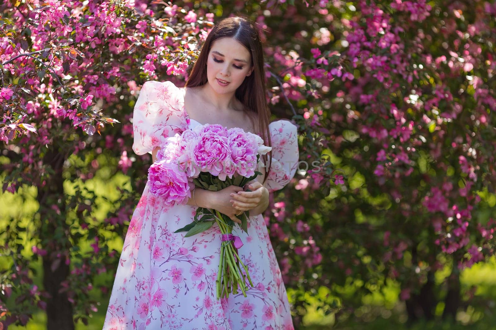 pretty brunette in a light pink dress, with a large bouquet of pink peonies, stands near pink blooming apple trees,in the garden on a sunny day. Copy space.