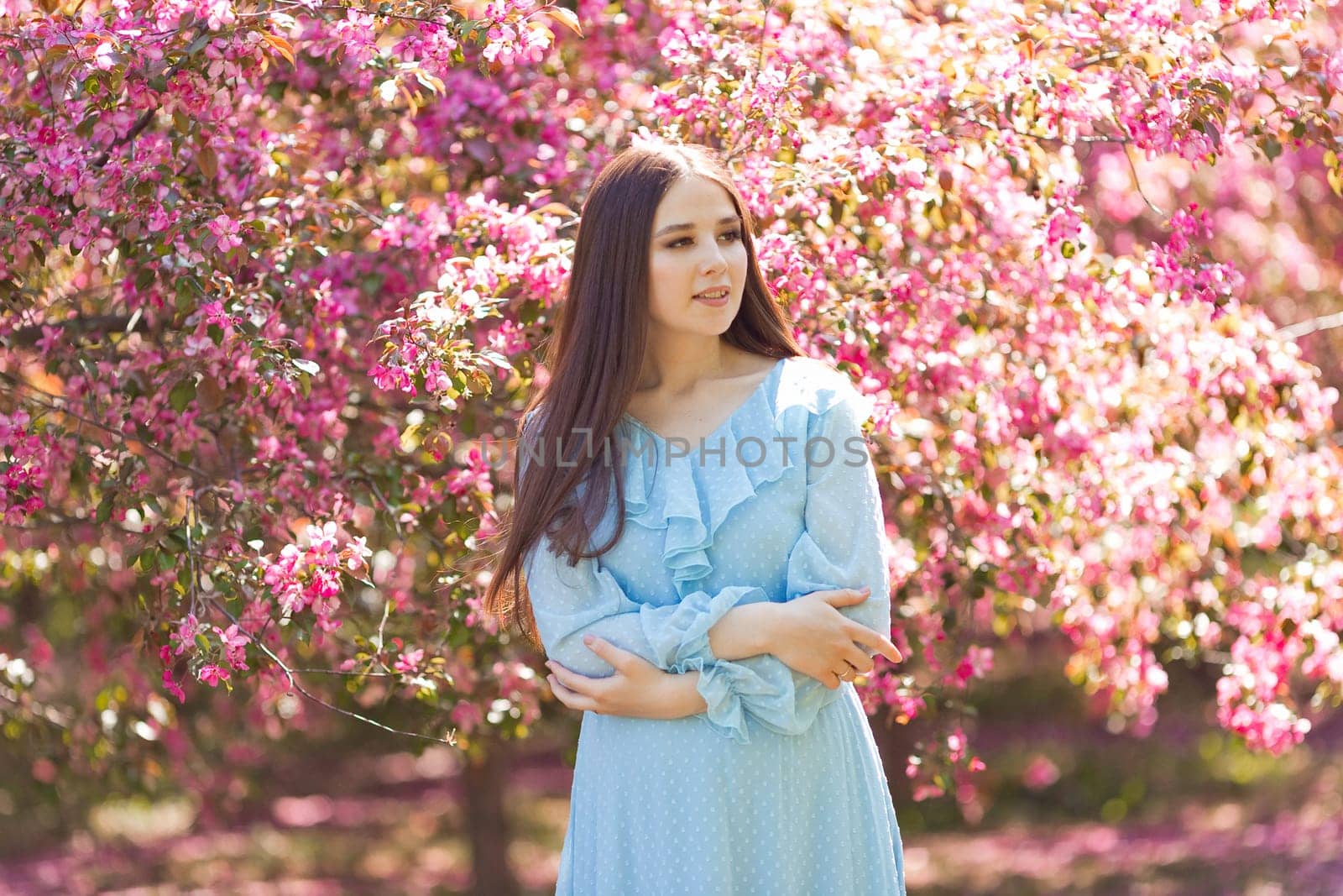 A smiling girl, in light blue dress, standing in a pink blooming garden, looking away. Close up. Copy space