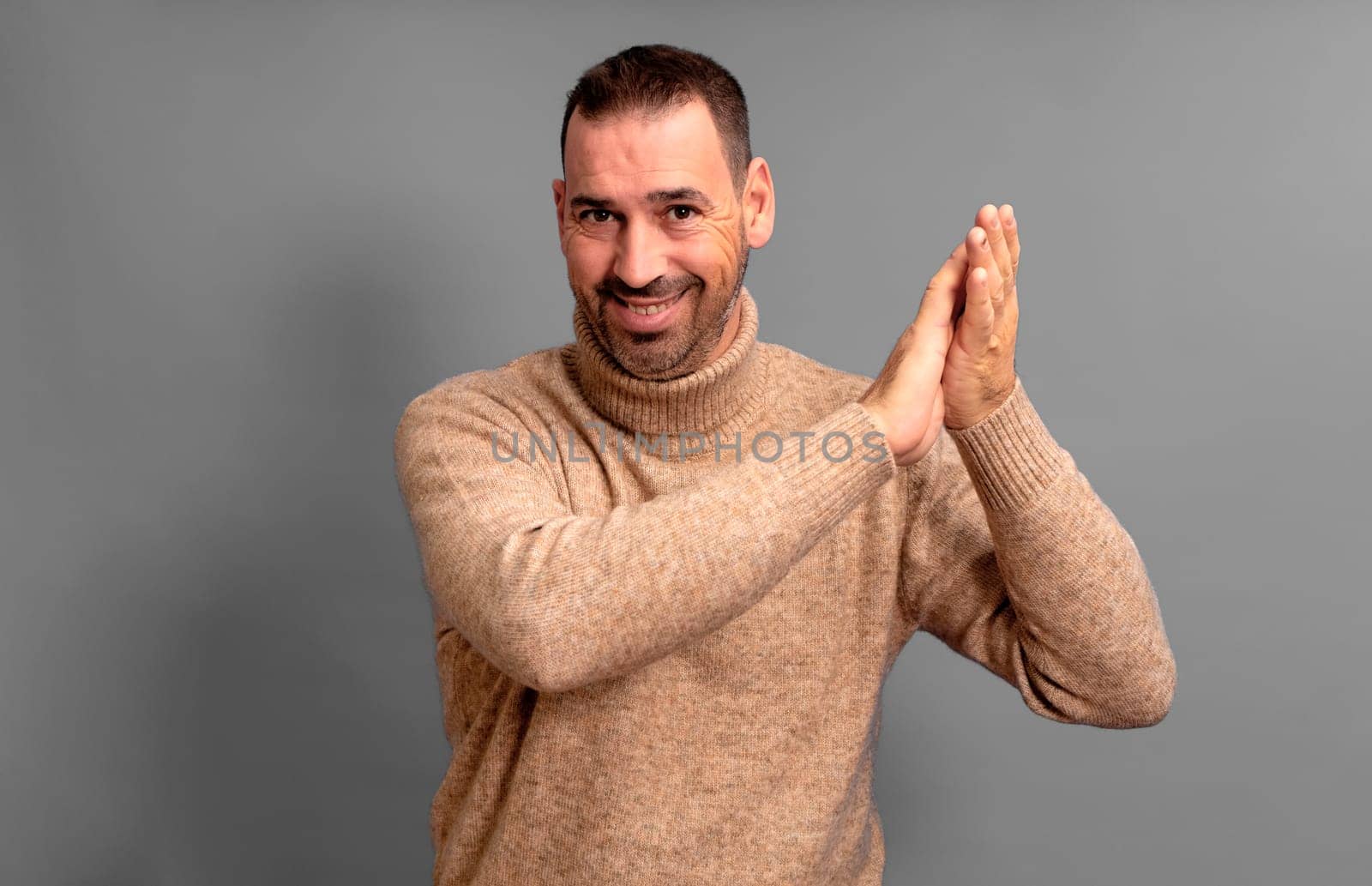 Bearded Hispanic man wearing a beige turtleneck clapping his hands proud and pleased at the spectacle he just witnessed, isolated over gray background. by Barriolo82