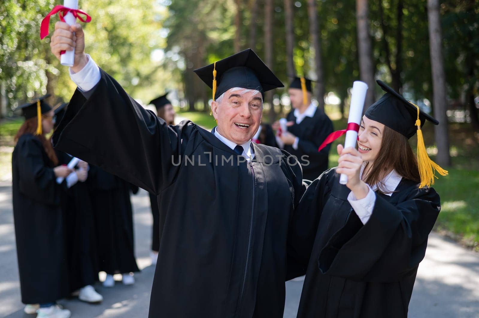 A group of graduates in robes outdoors. An elderly man and a young woman congratulate each other on their graduation