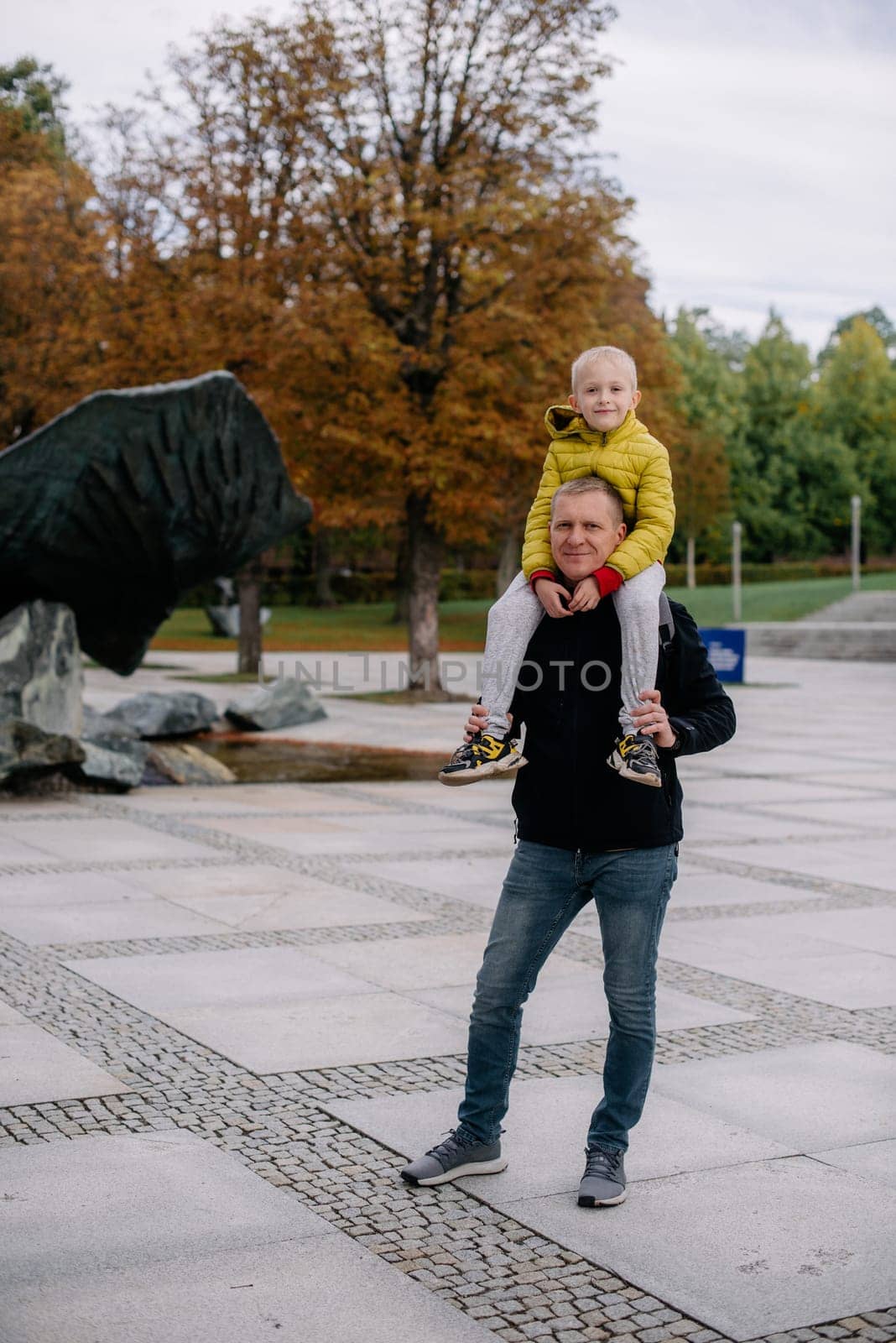 Dad holds on his son's shoulders. Beautiful family is spending time together outside. Dad and his little son are having fun on a roof terrace with view on a city. Sitting on father's shoulders and smiling.