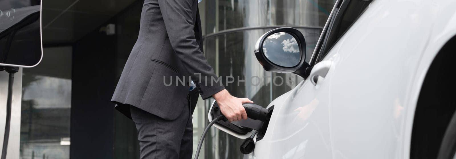 Below view closeup image of progressive black suit businessman recharge battery of his electric vehicle from public charging station. Renewable and alternative energy powered car concept.