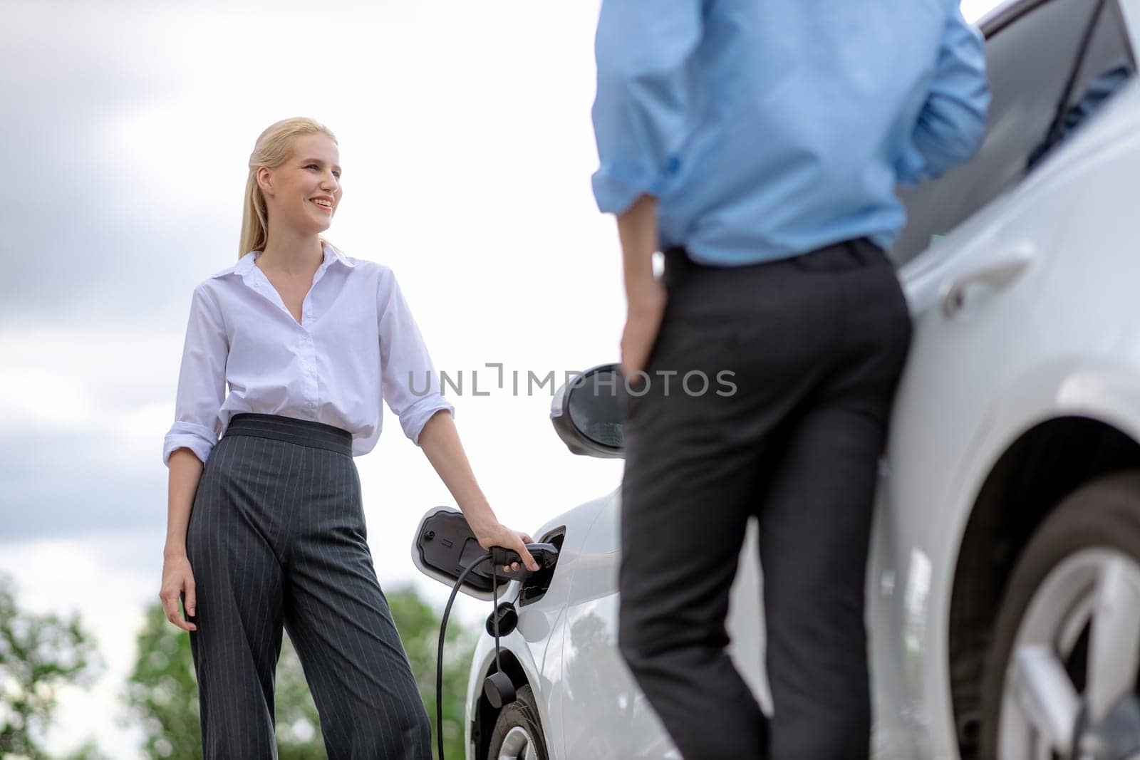 Progressive businessman and businesswoman with electric car parking and connected to public charging station before driving around city center. Eco friendly rechargeable car powered by clean energy.