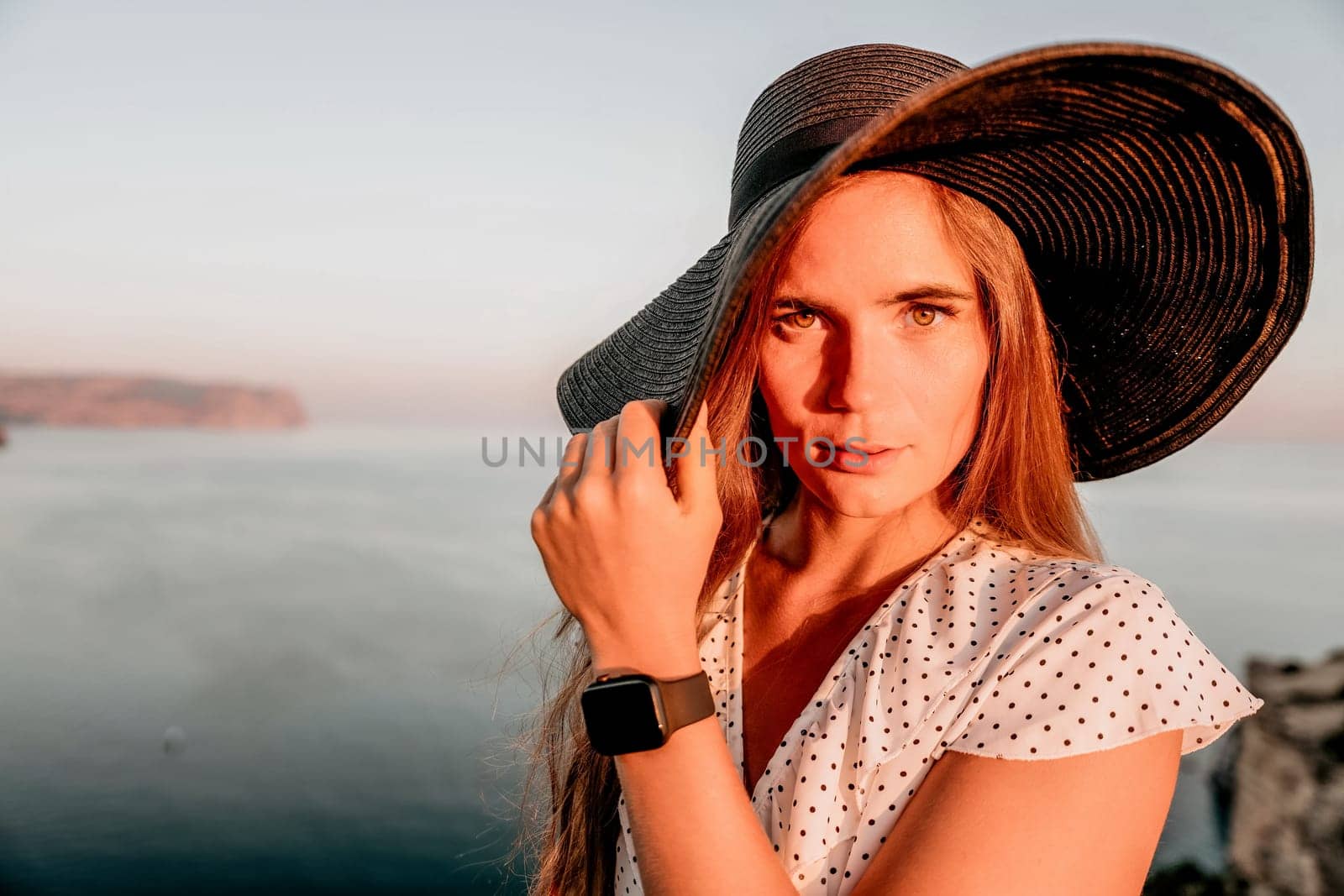 Portrait of happy young woman wearing summer black hat with large brim at beach on sunset. Closeup face of attractive girl with black straw hat. Happy young woman smiling and looking at camera at sea