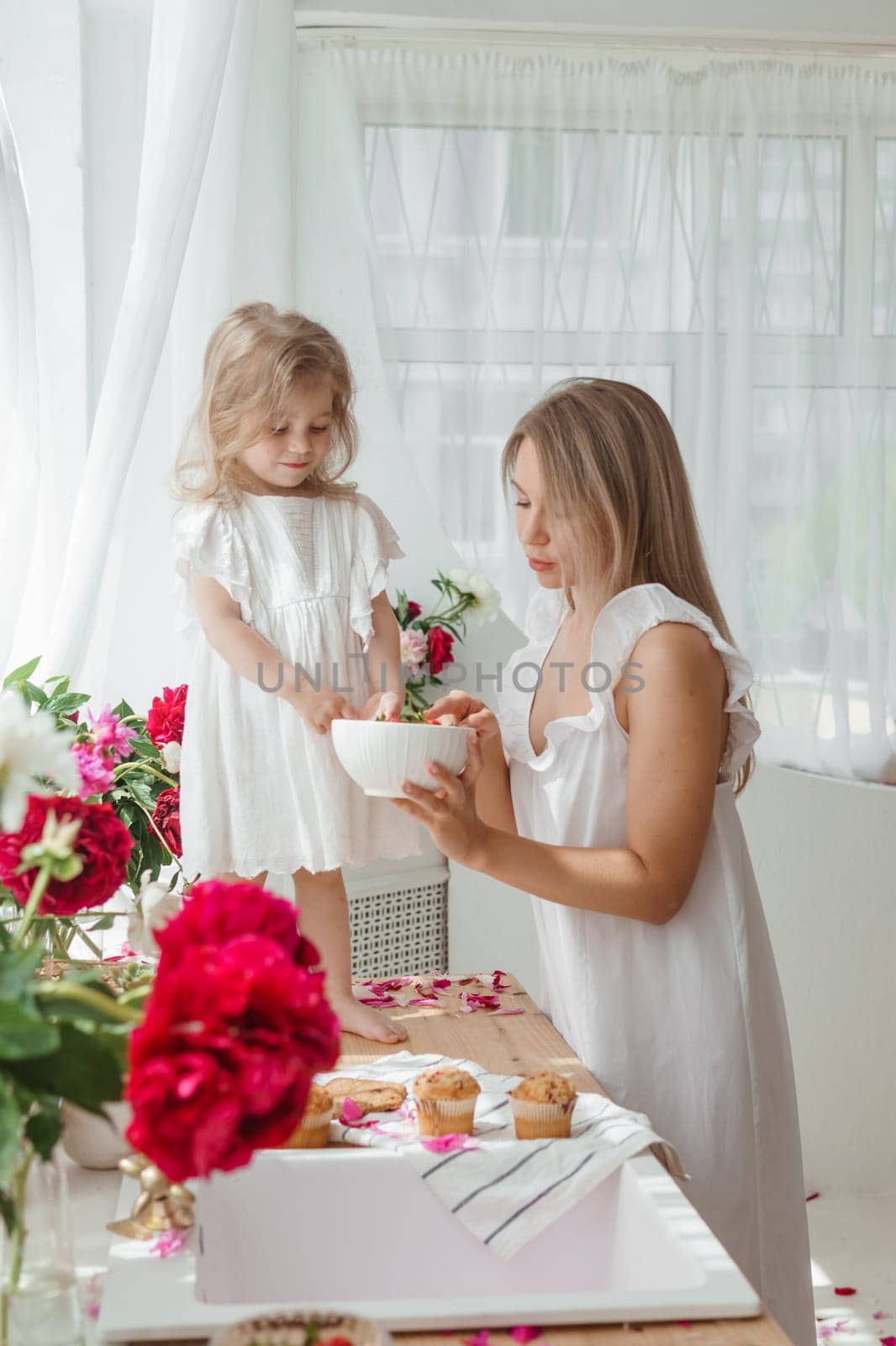 A little blonde girl with her mom on a kitchen countertop decorated with peonies. The concept of the relationship between mother and daughter. Spring atmosphere