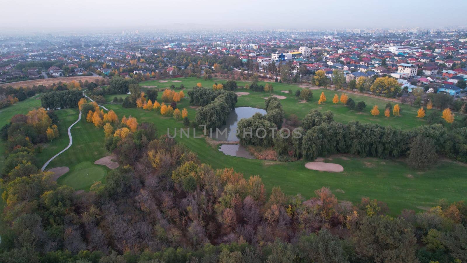 People play golf on a green field in autumn. Yellow-red leaves on some trees. Places for holes. There is a golf car. Houses and mountains are visible in the distance in a haze. Sunset. Wedding