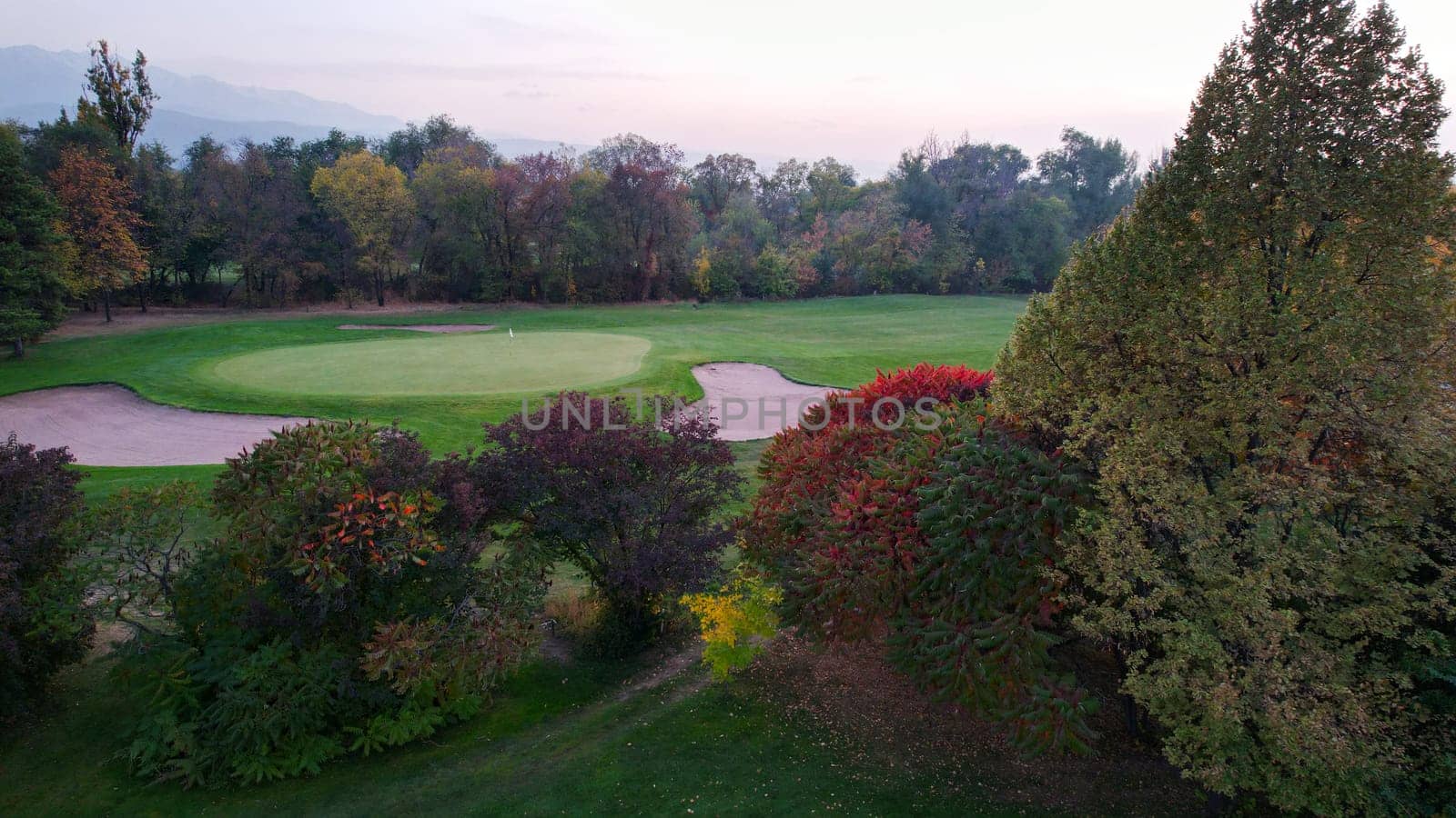 People play golf on a green field in autumn. Yellow-red leaves on some trees. Places for holes. There is a golf car. Houses and mountains are visible in the distance in a haze. Sunset. Wedding