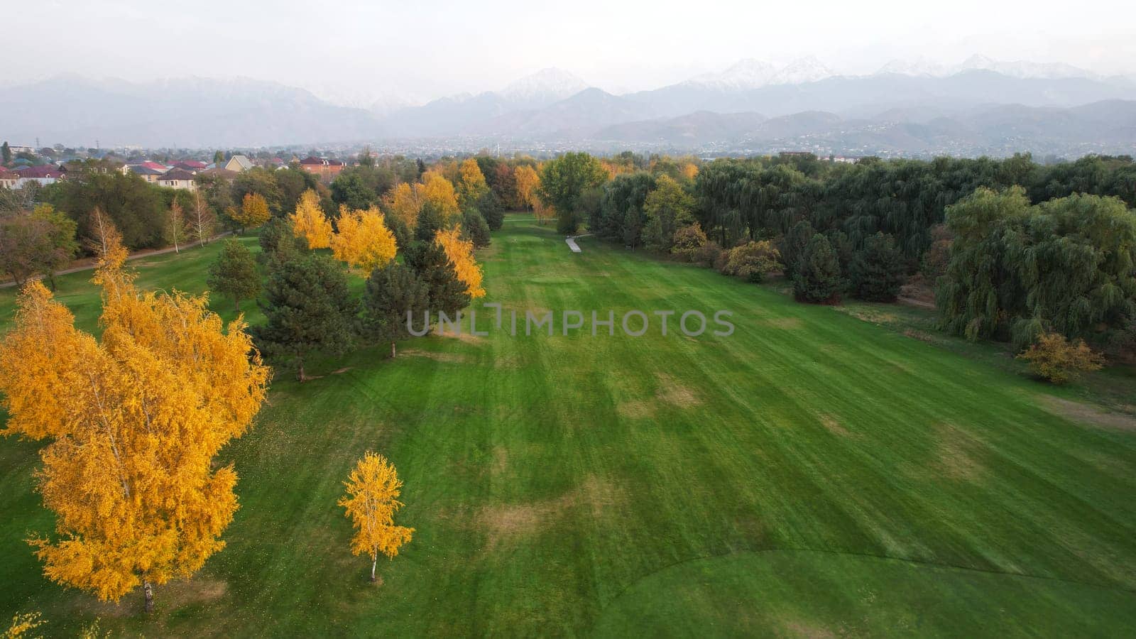 People play golf on a green field in autumn. Yellow-red leaves on some trees. Places for holes. There is a golf car. Houses and mountains are visible in the distance in a haze. Sunset. Wedding