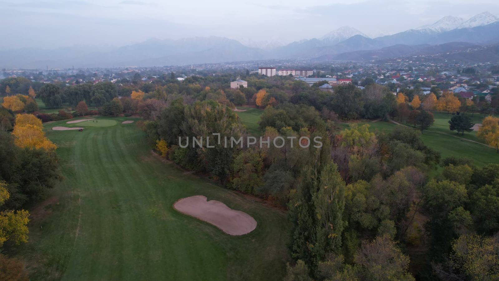 People play golf on a green field in autumn. Yellow-red leaves on some trees. Places for holes. There is a golf car. Houses and mountains are visible in the distance in a haze. Sunset. Wedding