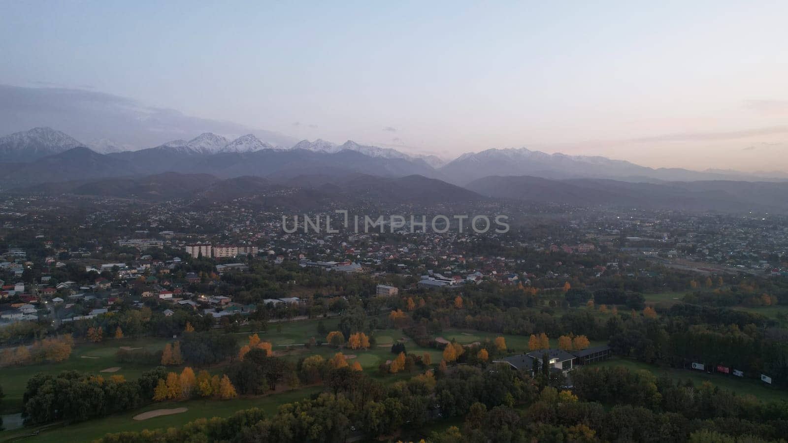 People play golf on a green field in autumn. Yellow-red leaves on some trees. Places for holes. There is a golf car. Houses and mountains are visible in the distance in a haze. Sunset. Wedding