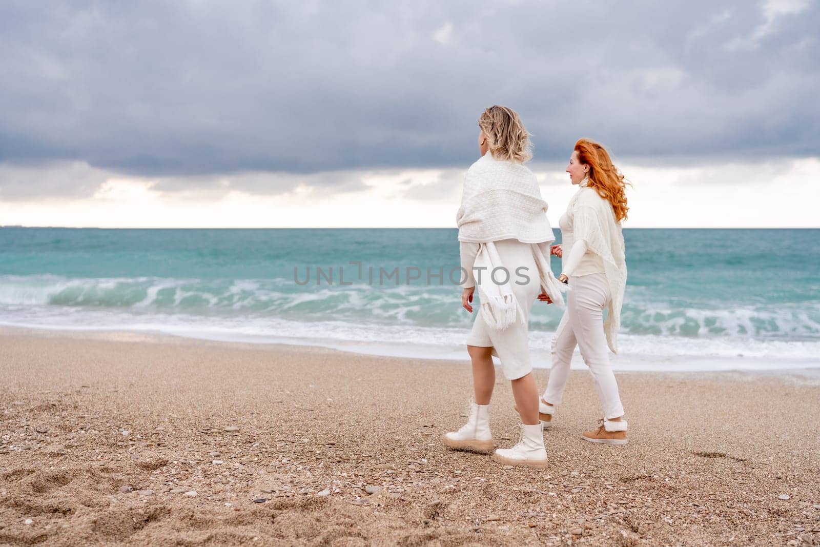 Women sea walk friendship spring. Two girlfriends, redhead and blonde, middle-aged walk along the sandy beach of the sea, dressed in white clothes. Against the backdrop of a cloudy sky and the winter sea. Weekend concept