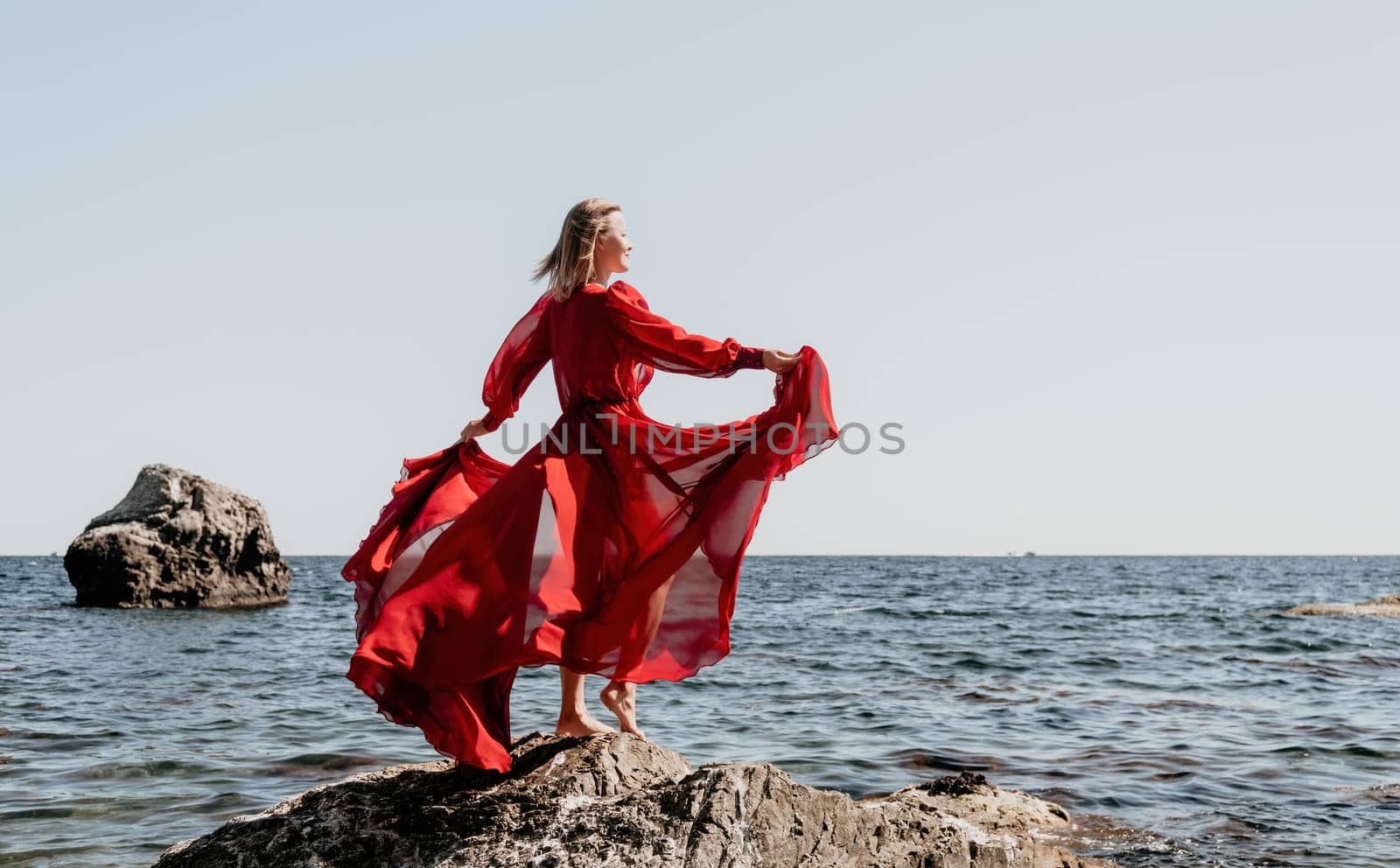 Woman travel sea. Young Happy woman in a long red dress posing on a beach near the sea on background of volcanic rocks, like in Iceland, sharing travel adventure journey