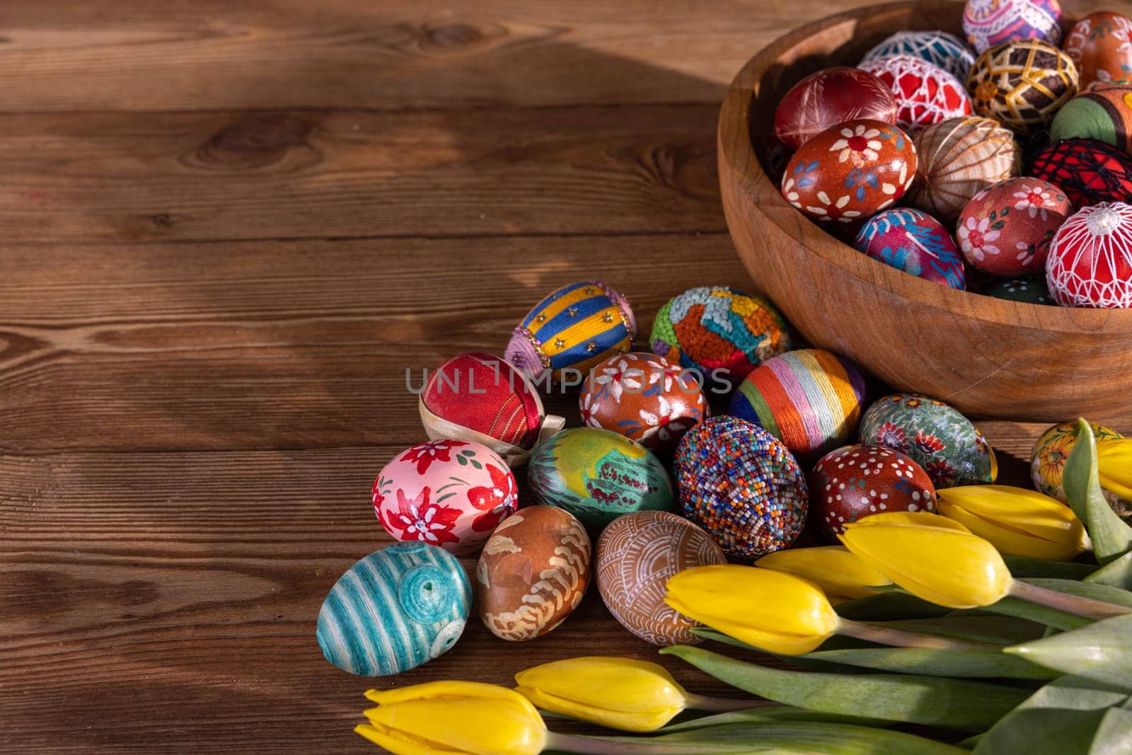 Intricately arranged Easter eggs on a wooden tabletop and in a wooden bowl. The Easter composition is completed with swishy yellow tulips.