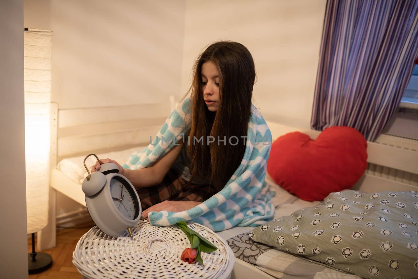 A teenage girl takes a large alarm clock from the cabinet. A girl under a soft blanket. The light of a lamp on the wall.
