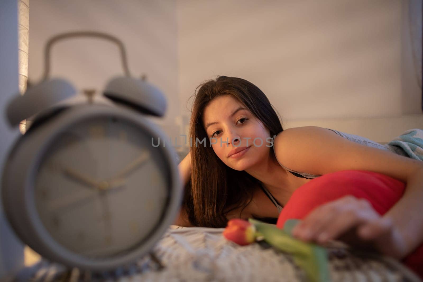 The brunette reaches for a flower on the nightstand. The light of a lamp on the wall. A large alarm clock stands on the bedside table.