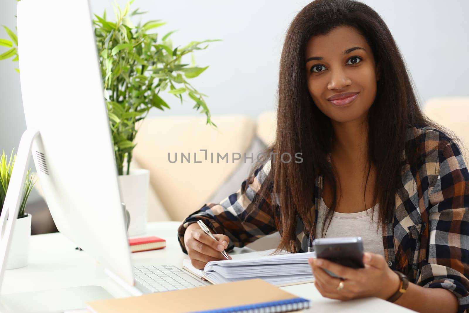Smiling african american woman holding phone making notes in notebook checks, writes down important information. Afro business woman planning working day in office concept