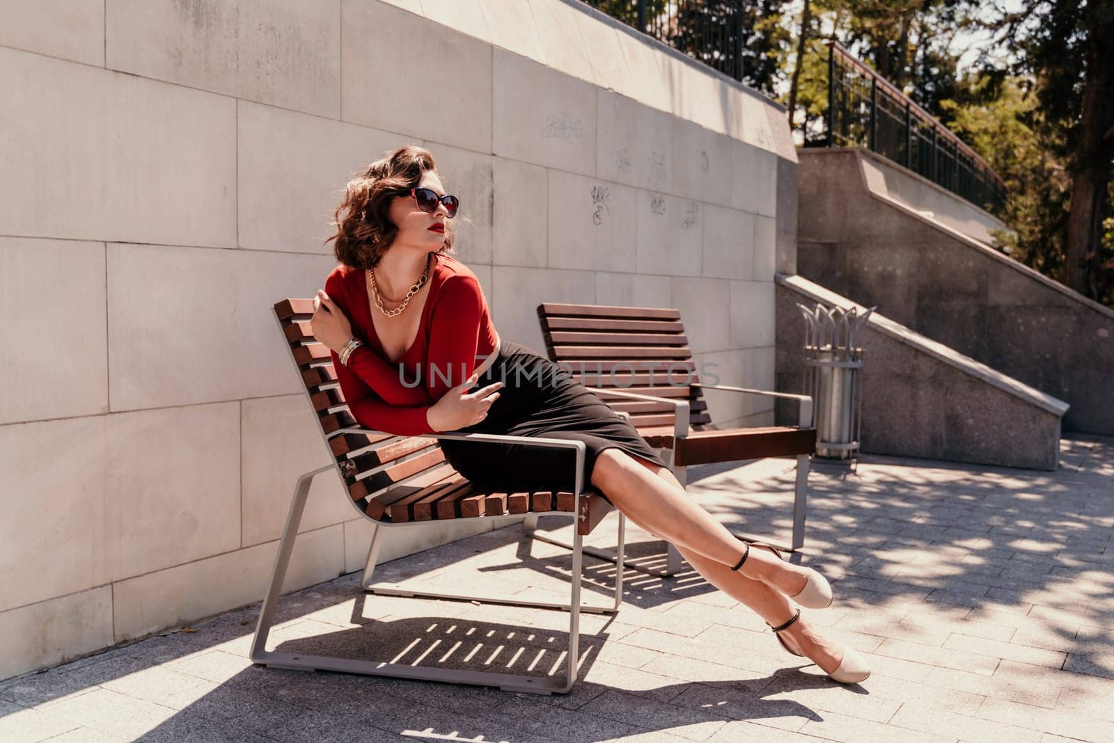 Portrait of a woman on the street. An attractive woman in glasses, a red blouse and a black skirt is sitting on a bench outside