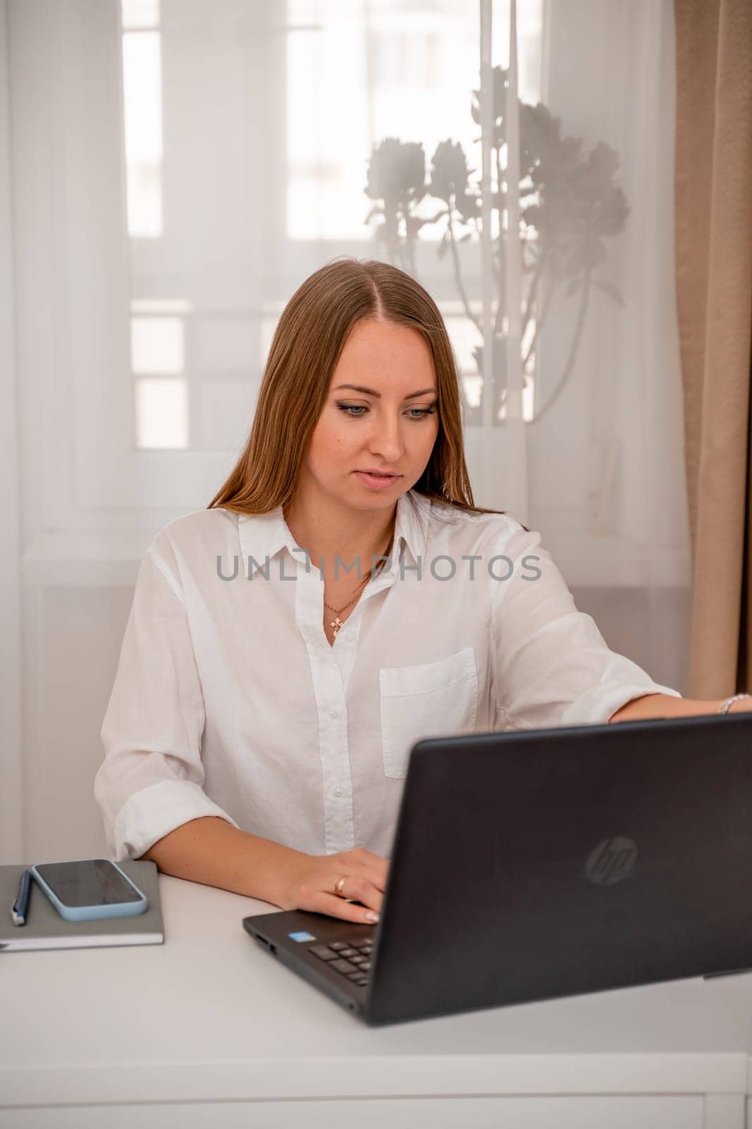 European professional woman is sitting with a laptop at a table in a home office, a positive woman is studying while working on a PC. She is wearing a beige jacket and jeans and is on the phone. by Matiunina