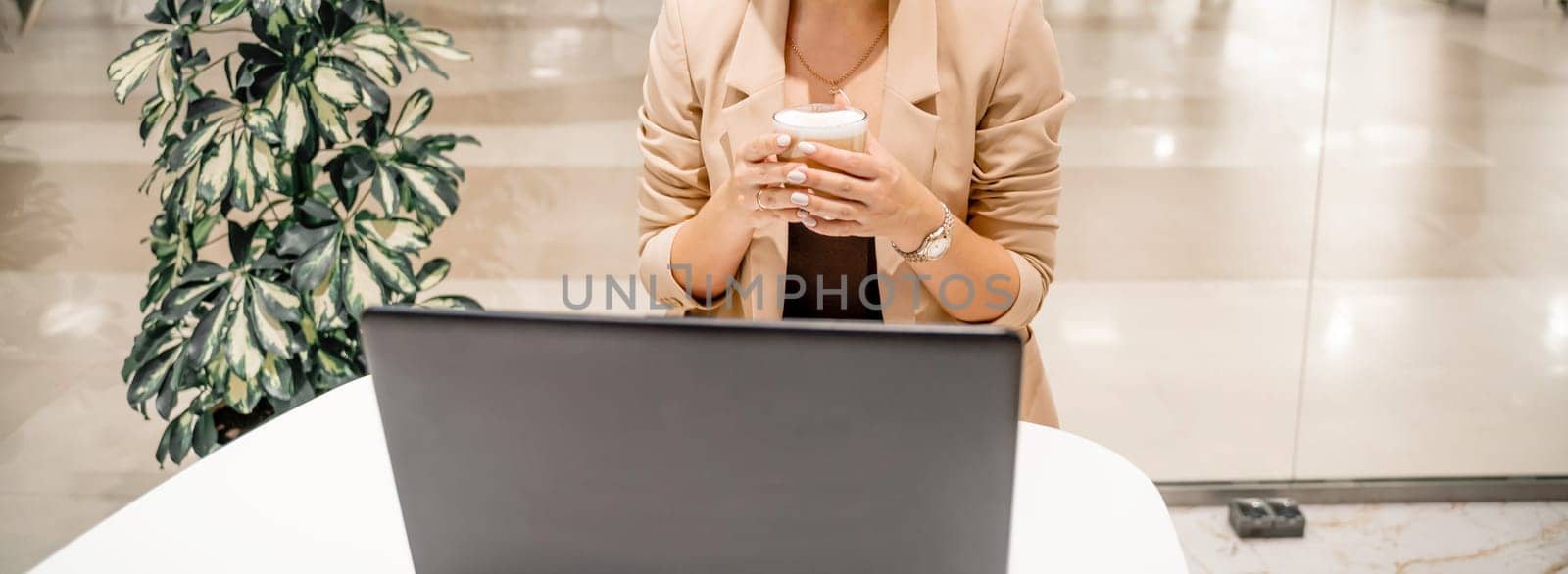 A business woman sits in a cafe, works at a computer, drinks coffee. She is wearing a beige jacket, brown top and black trousers. by Matiunina