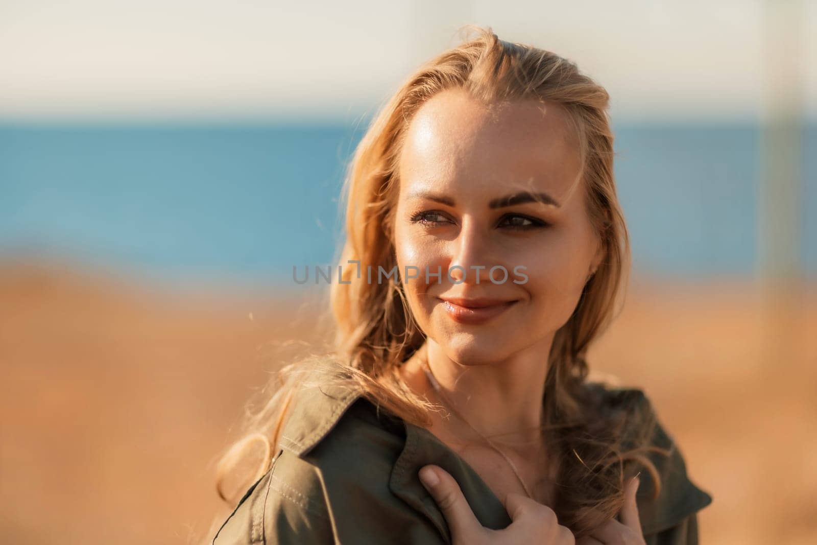 Portrait blonde sea cape. A calm young blonde in an unbuttoned khaki raincoat stands on the seashore, under the raincoat there is a black skirt and top.