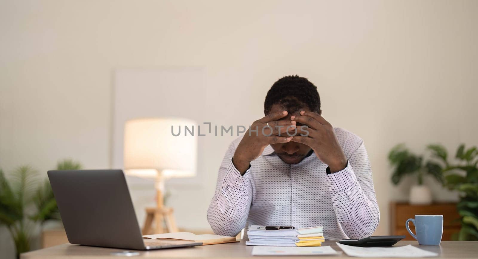 Concentrated business african american man working from home, sitting at work desk, using laptop, looking at laptop thoughtfully, hands near face, thinking about financial business plan or new startup by wichayada