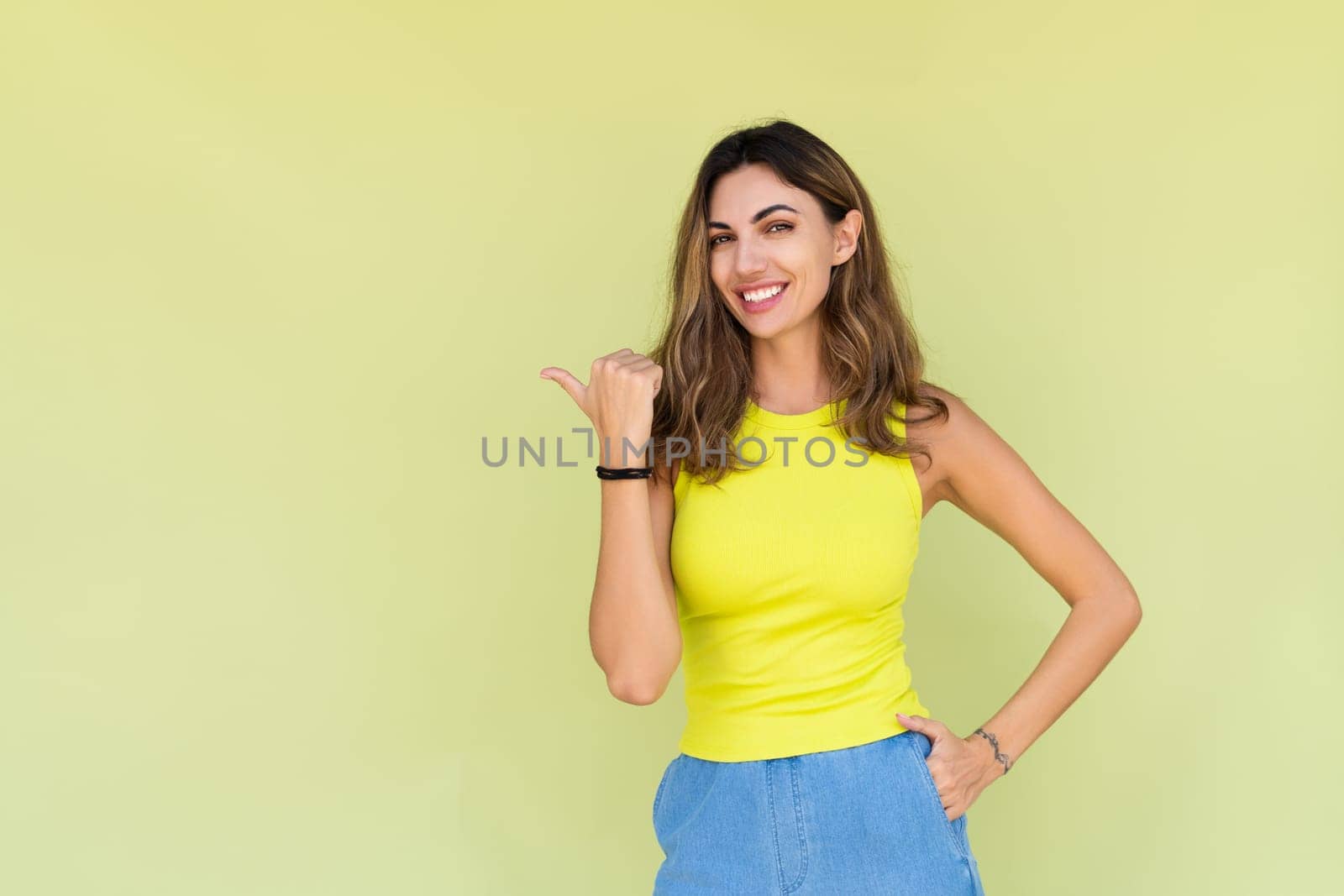 Young brunette in casual wear isolated on green background happy look to camera smile excited point finger to left