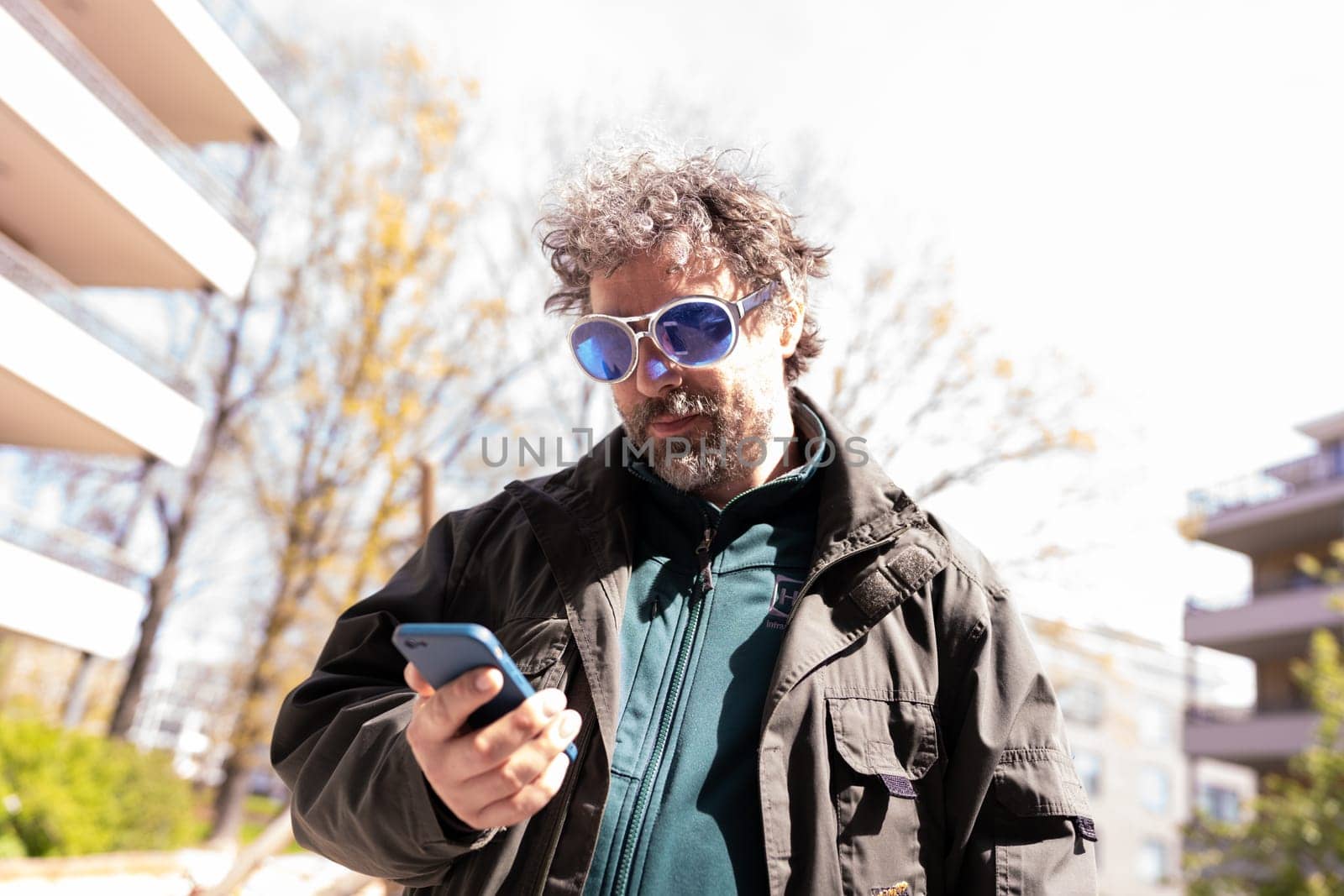 Mature man with grey hair in blue sunglasses looking at his smartphone by Varaksina