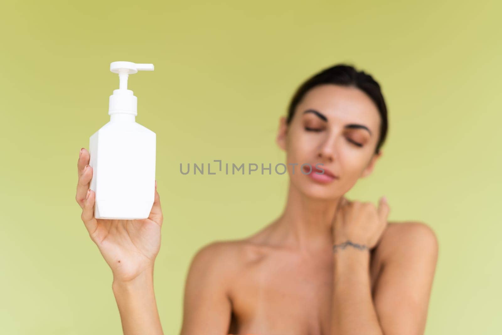 Beauty portrait of young topless woman with bare shoulders on green background with perfect skin and natural makeup holding bottle of shampoo, body lotion