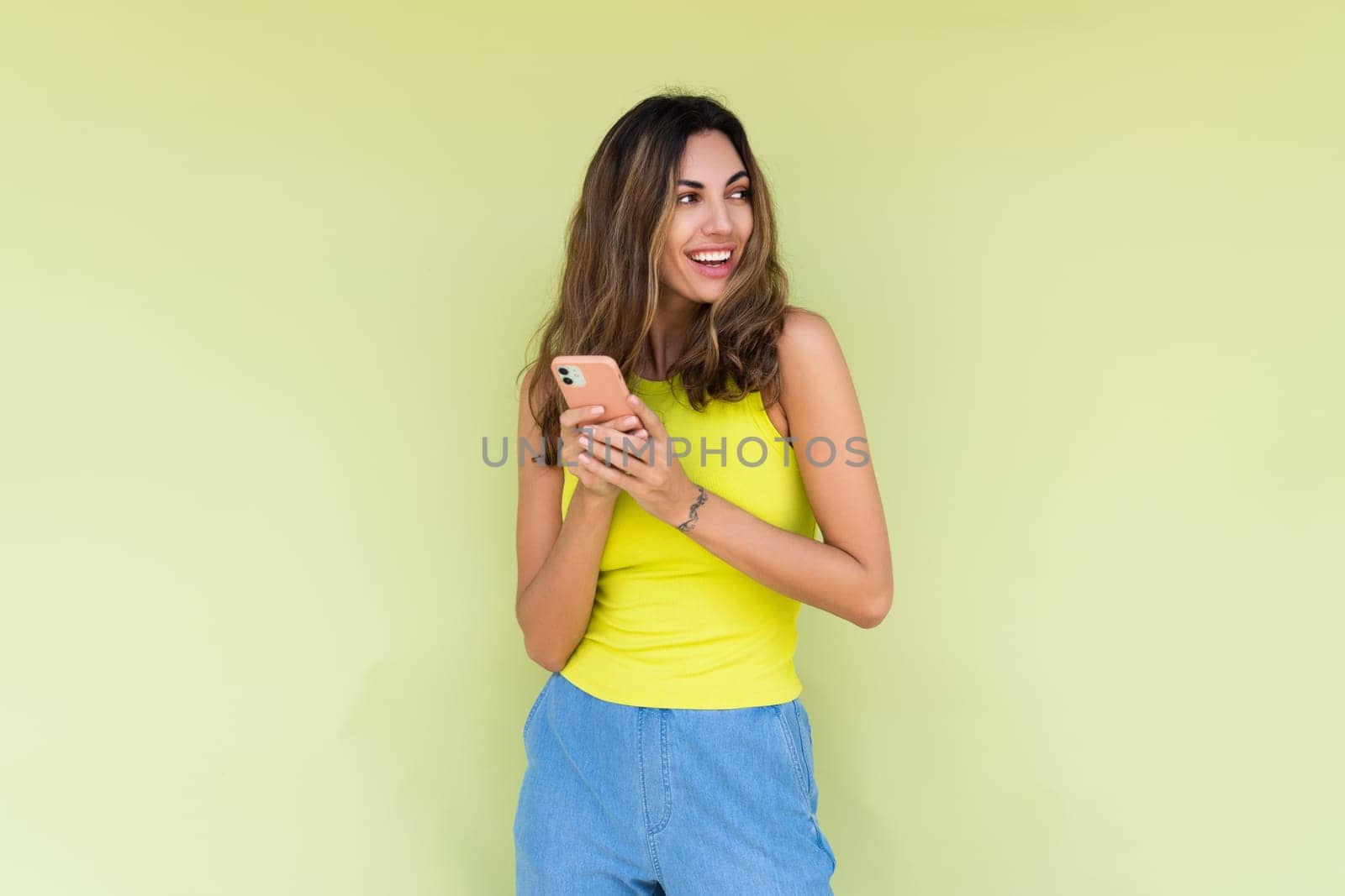 Young woman in casual wear isolated on green background holding phone and look aside with big smile