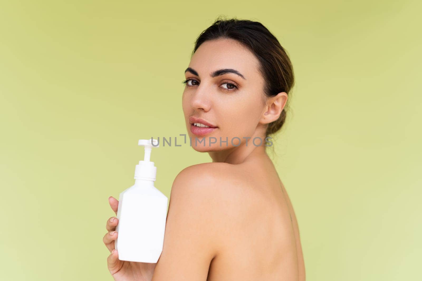 Beauty portrait of young topless woman with bare shoulders on green background with perfect skin and natural makeup holding bottle of shampoo, body lotion