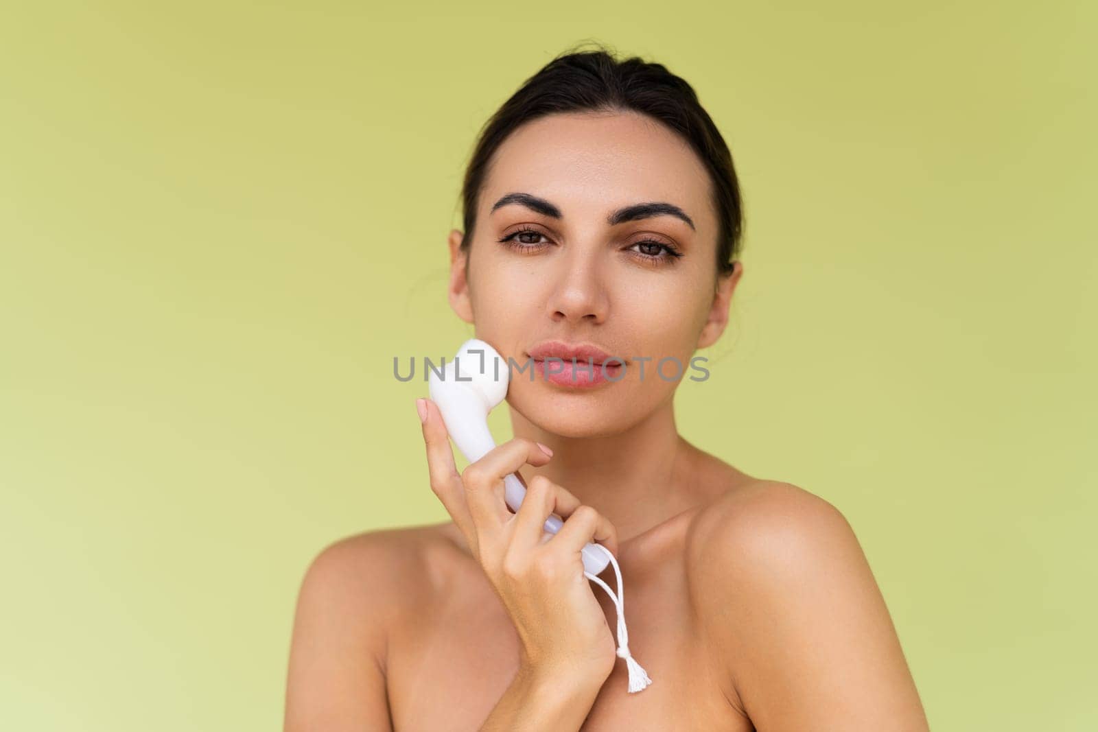 Beauty portrait of young topless woman with bare shoulders on green background with perfect skin and natural makeup holds face cleansing brush by kroshka_nastya