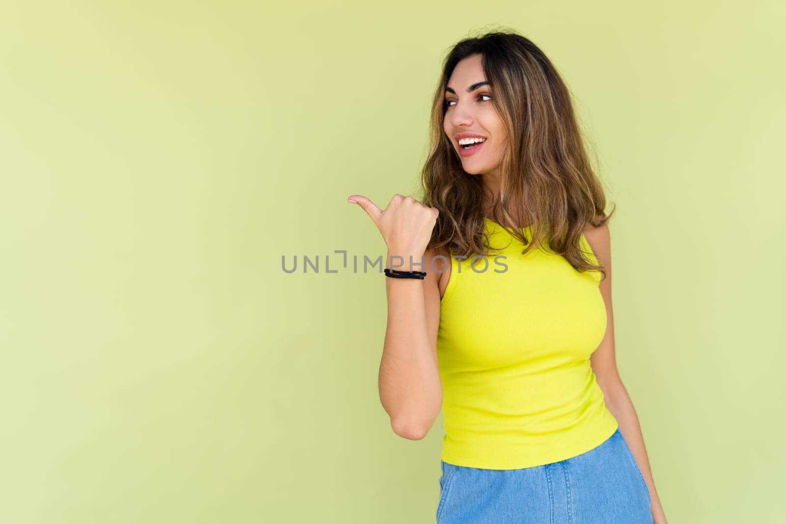 Young brunette in casual wear isolated on green background happy look to camera smile excited point thumb finger to left