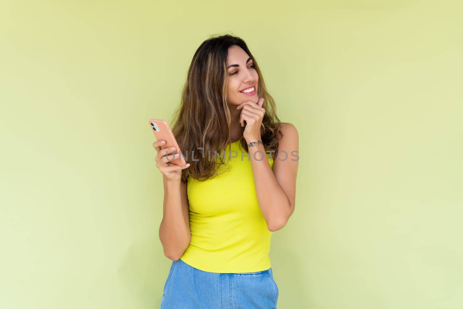 Young woman in casual wear isolated on green background holding phone and look aside with big thoughtful smile