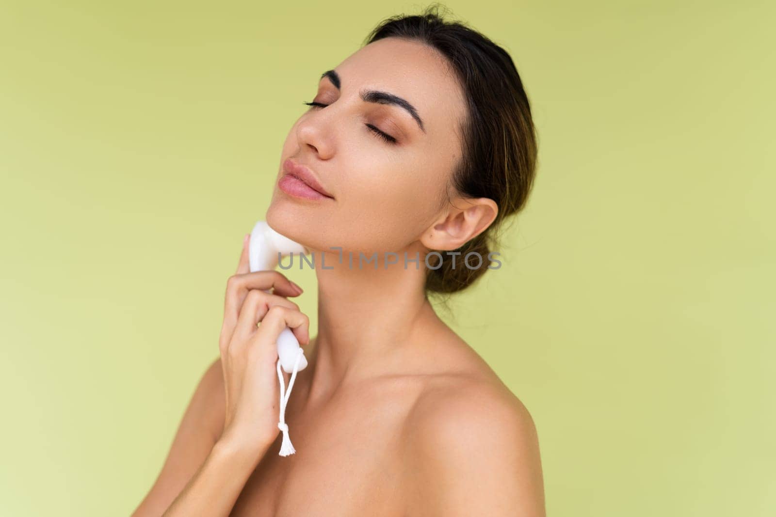 Beauty portrait of young topless woman with bare shoulders on green background with perfect skin and natural makeup holds face cleansing brush by kroshka_nastya