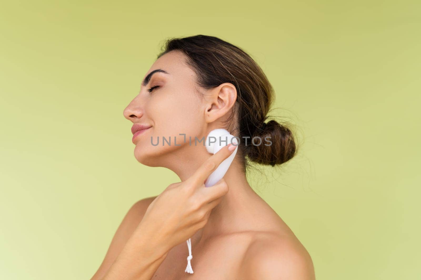 Beauty portrait of young topless woman with bare shoulders on green background with perfect skin and natural makeup holds face cleansing brush