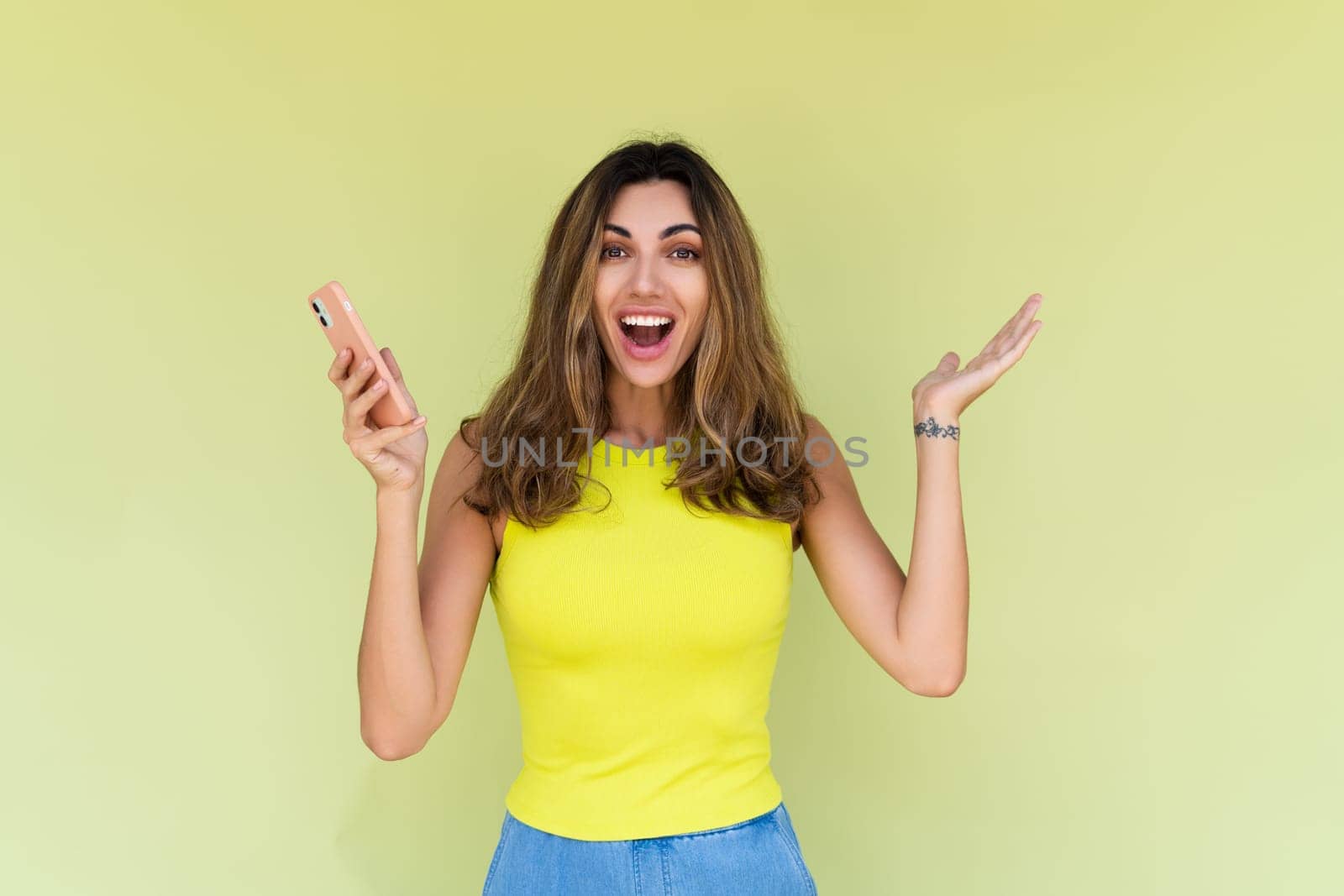 Young woman in casual wear isolated on green background holding phone and look to camera shocked amazed and surprised by kroshka_nastya