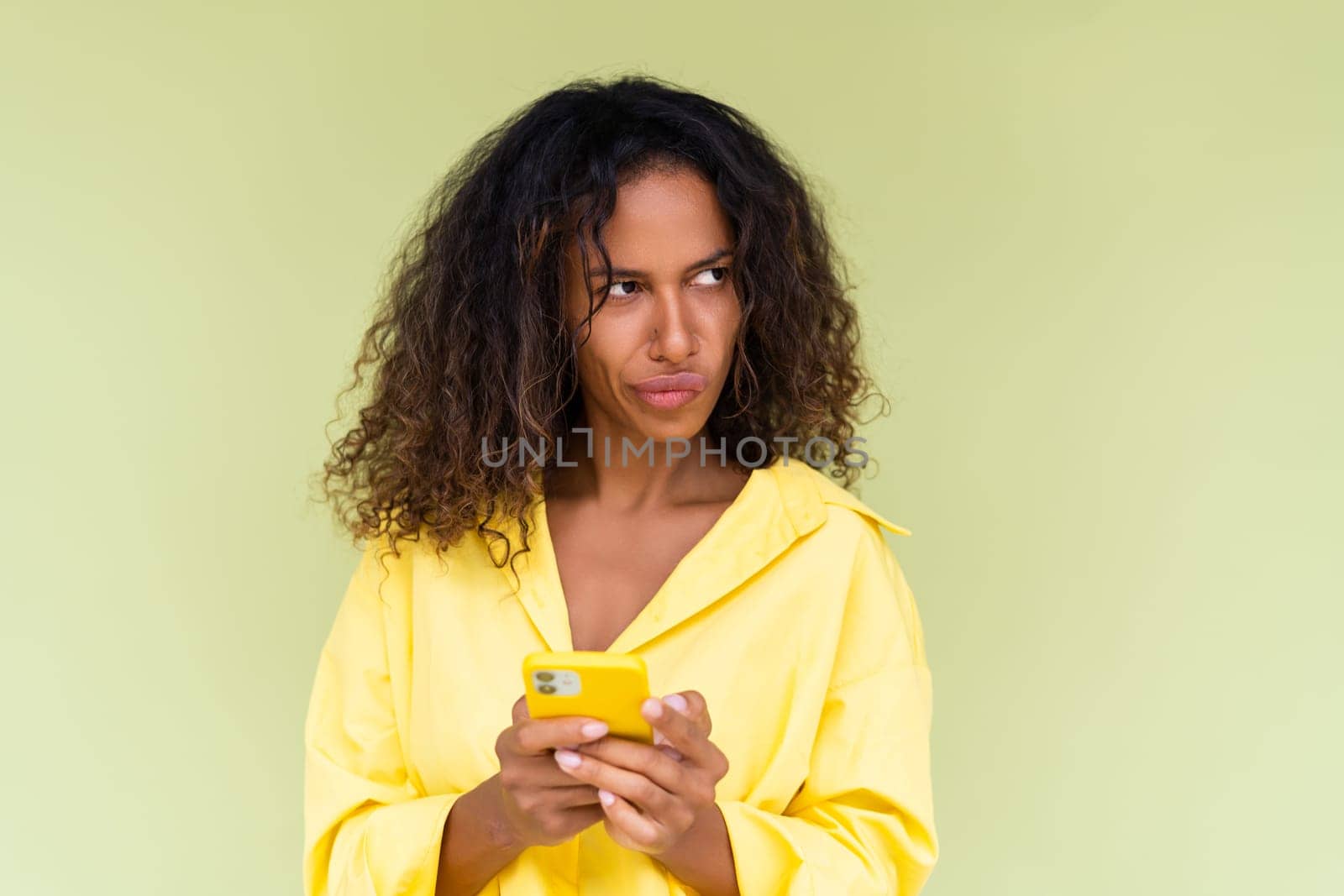 Beautiful african american woman in casual shirt on green background holds a phone confused thoughtful by kroshka_nastya