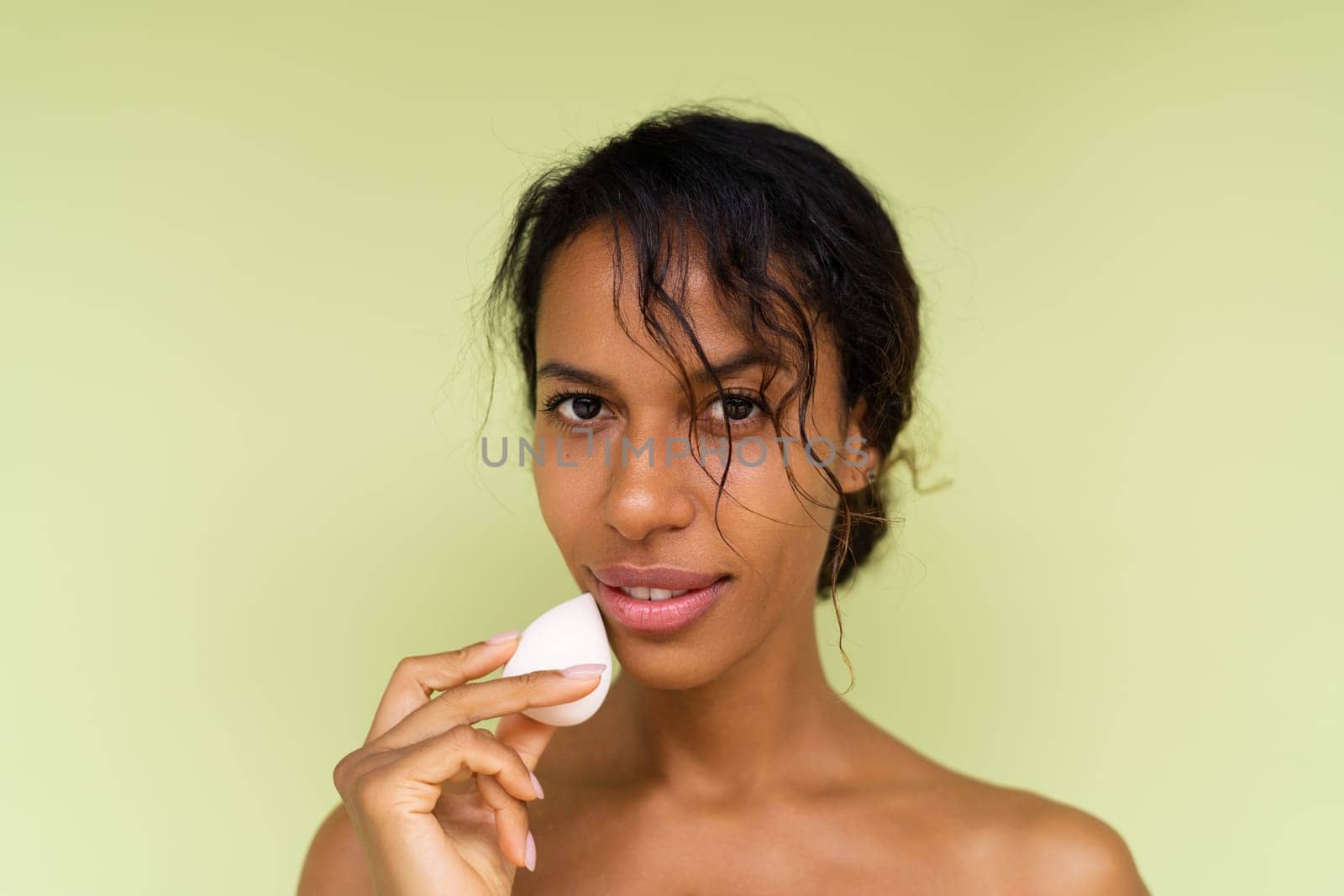 Beauty portrait of young topless african american woman with bare shoulders on green background with makeup foundation sponge