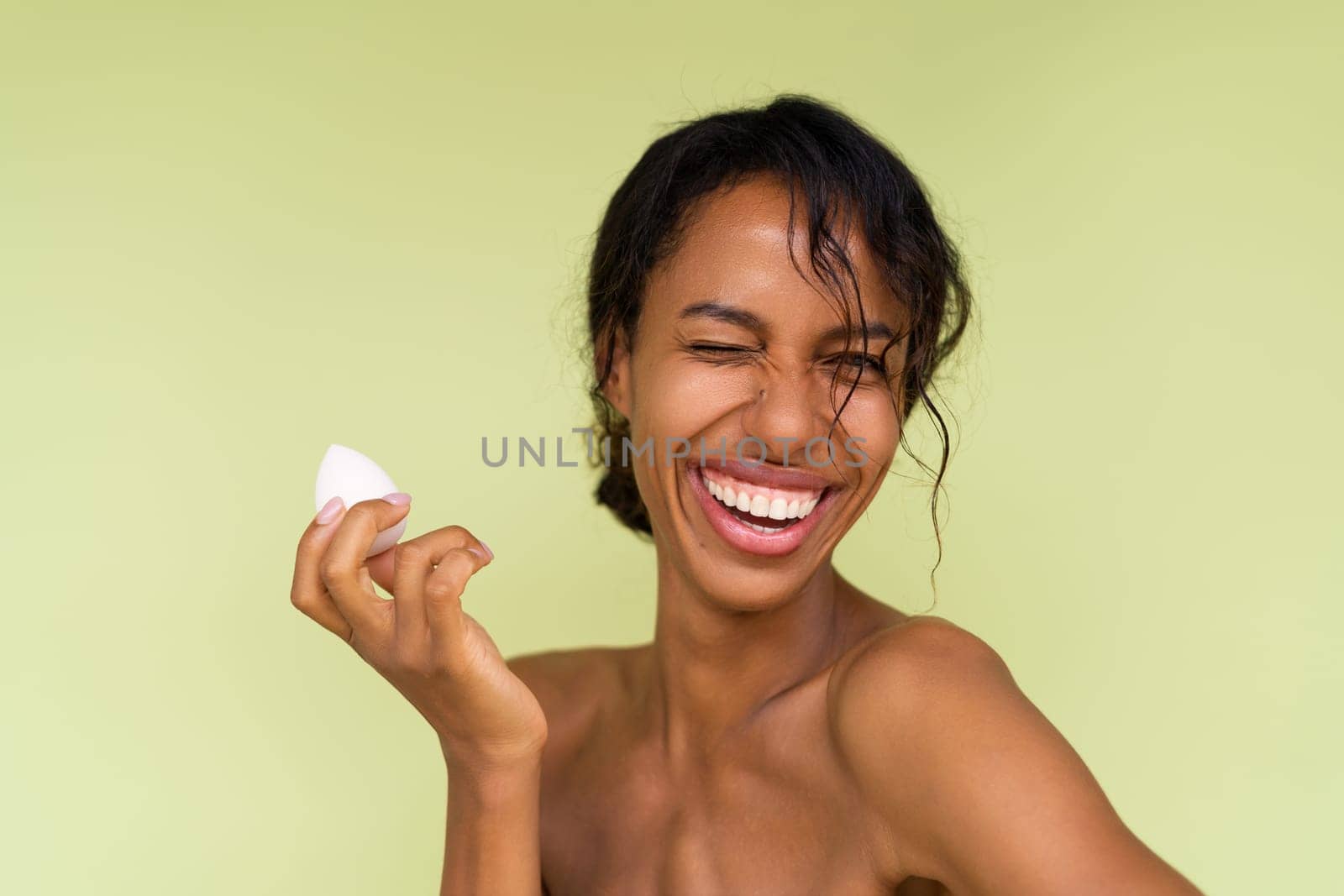 Beauty portrait of young topless african american woman with bare shoulders on green background with makeup foundation sponge