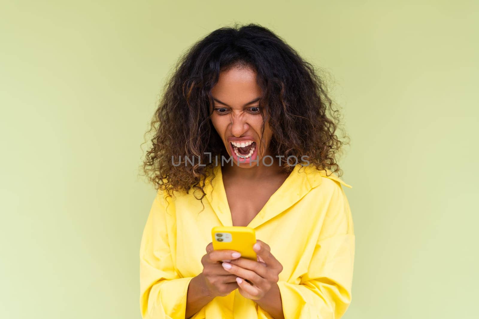 Beautiful african american woman in casual shirt on green background holds a phone look at screen with anger