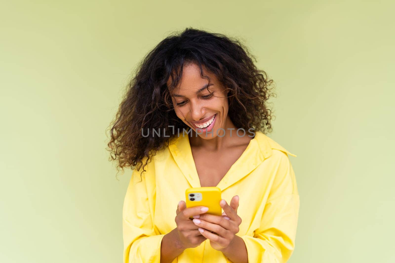 Beautiful african american woman in casual shirt on green background holds mobile phone with a smile