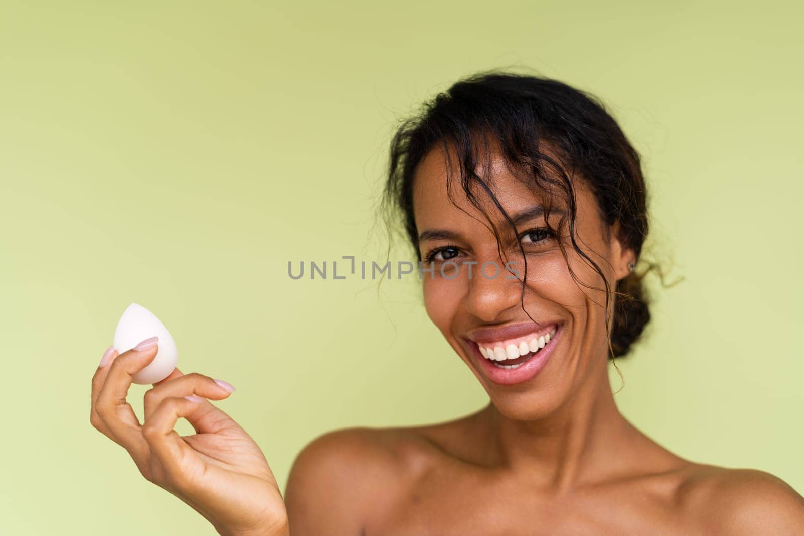 Beauty portrait of young topless african american woman with bare shoulders on green background with makeup foundation sponge
