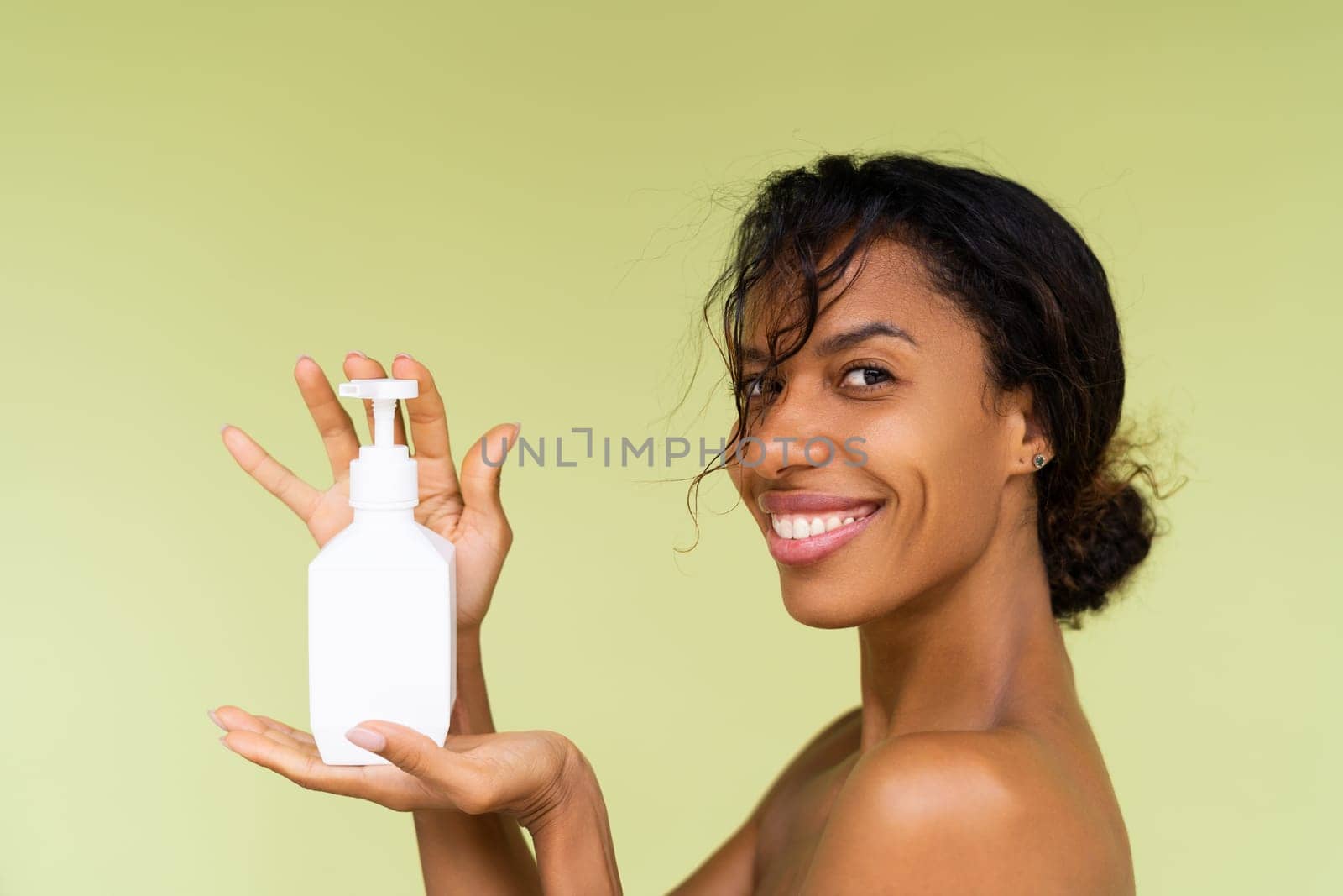Beauty portrait of young topless african american woman with bare shoulders on green background with white bottle of body lotion by kroshka_nastya
